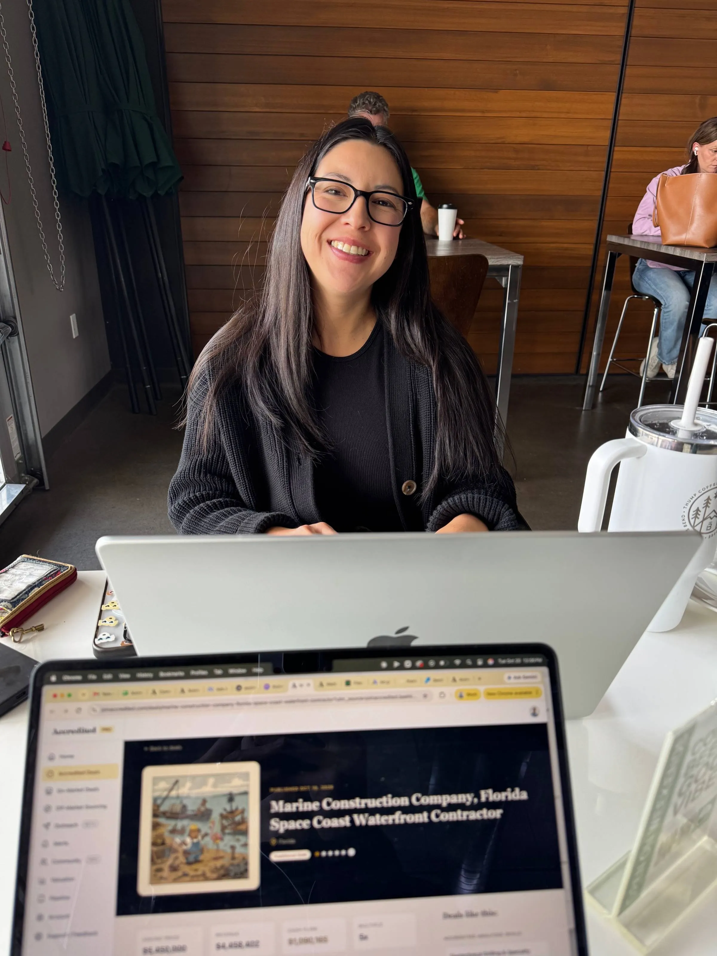 Ben’s wife smiling behind her laptop at a coffee shop table, with Ben’s laptop in the foreground