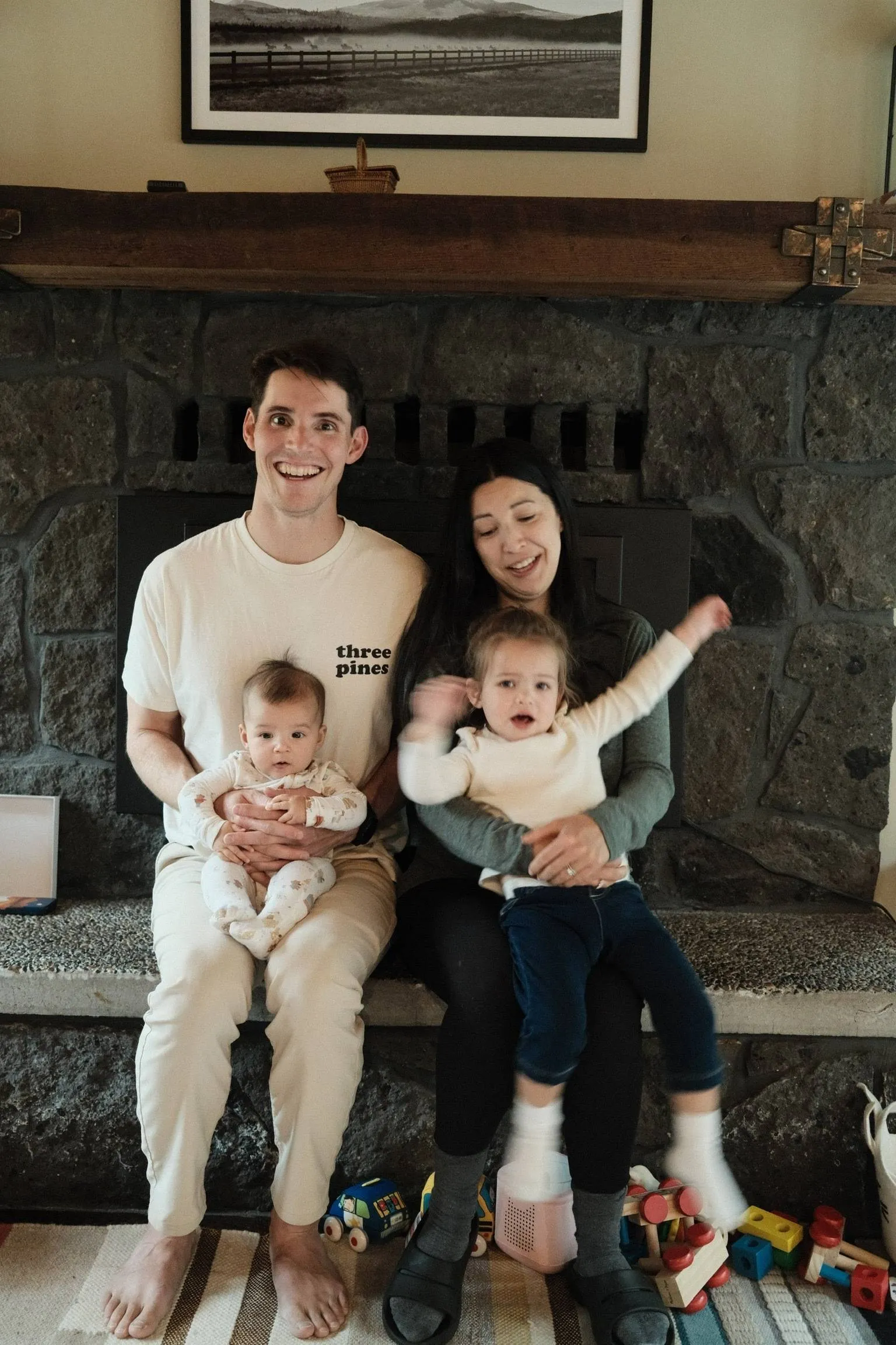Ben and his wife sitting on a stone fireplace hearth, each holding one of their kids, with toys scattered on the rug in front of them