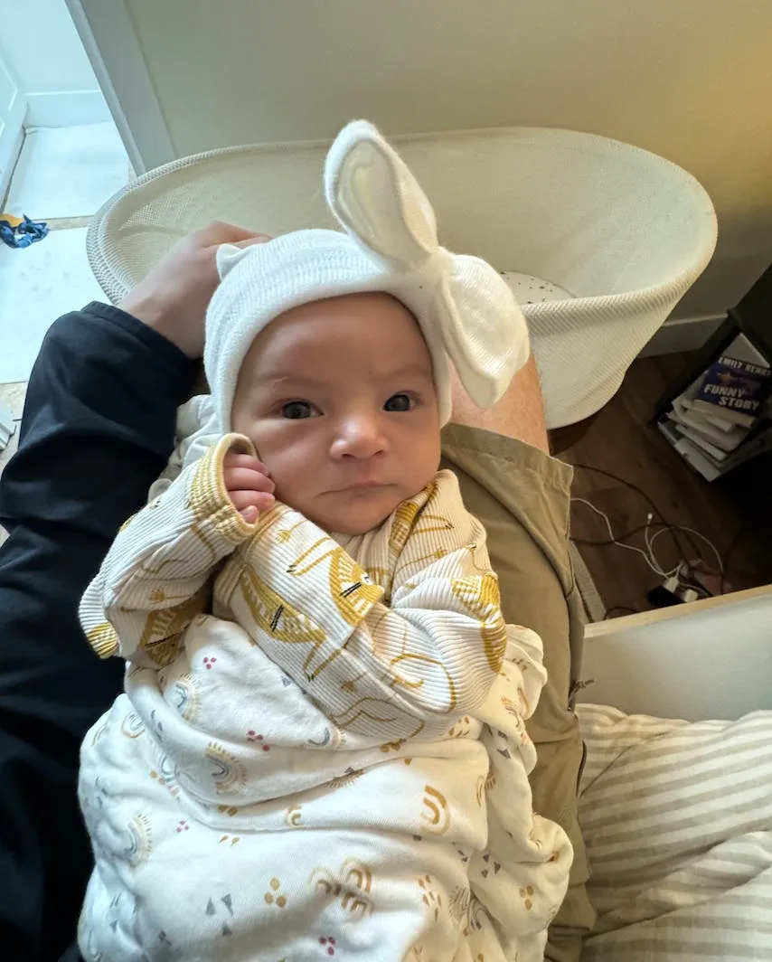 Newborn baby girl wearing a white bow headband, swaddled in a patterned blanket, resting on a parent’s lap beside a bassinet