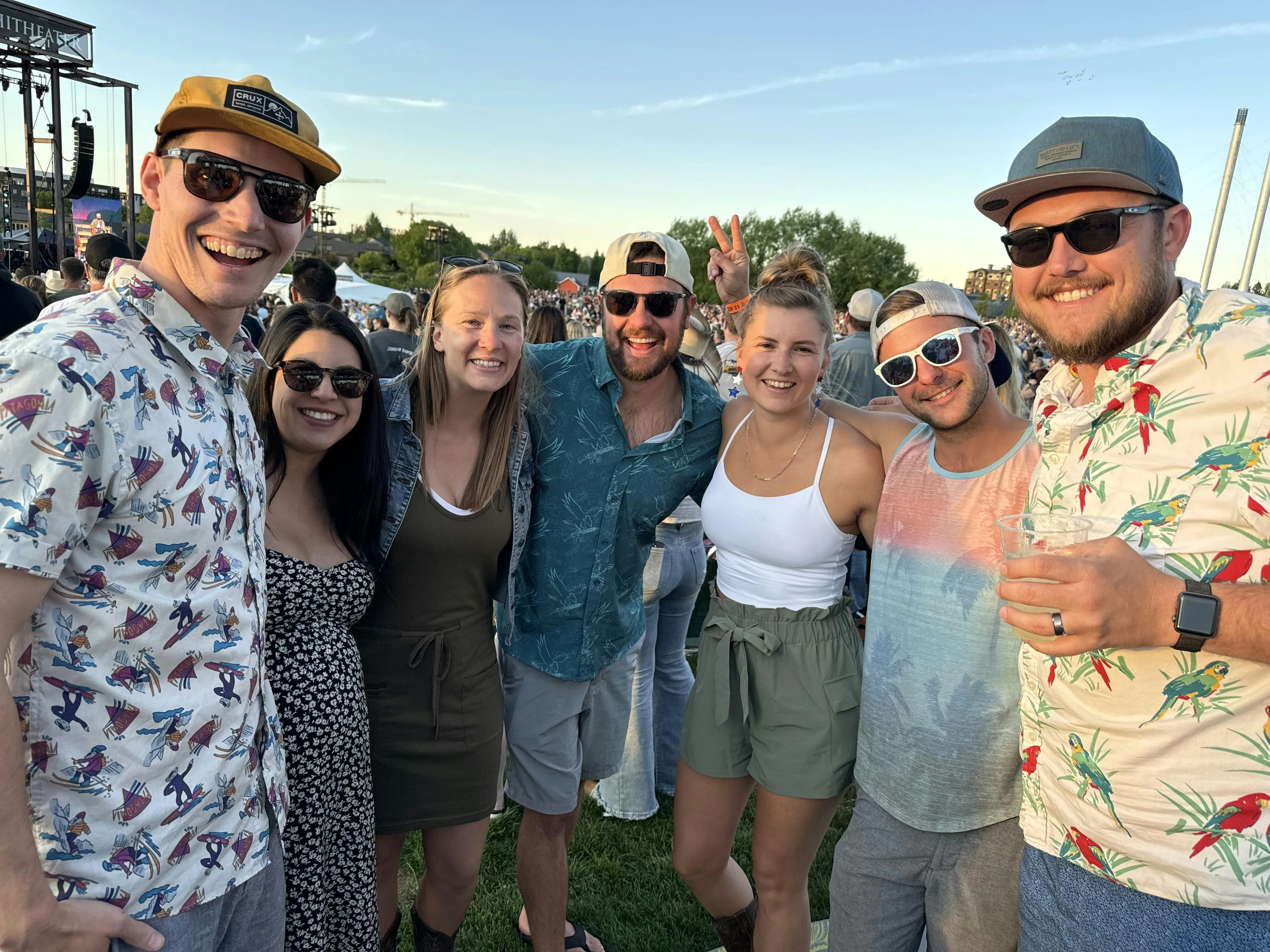 A group of seven friends in sunglasses and summer clothes smiling arm-in-arm at an outdoor amphitheater concert at sunset