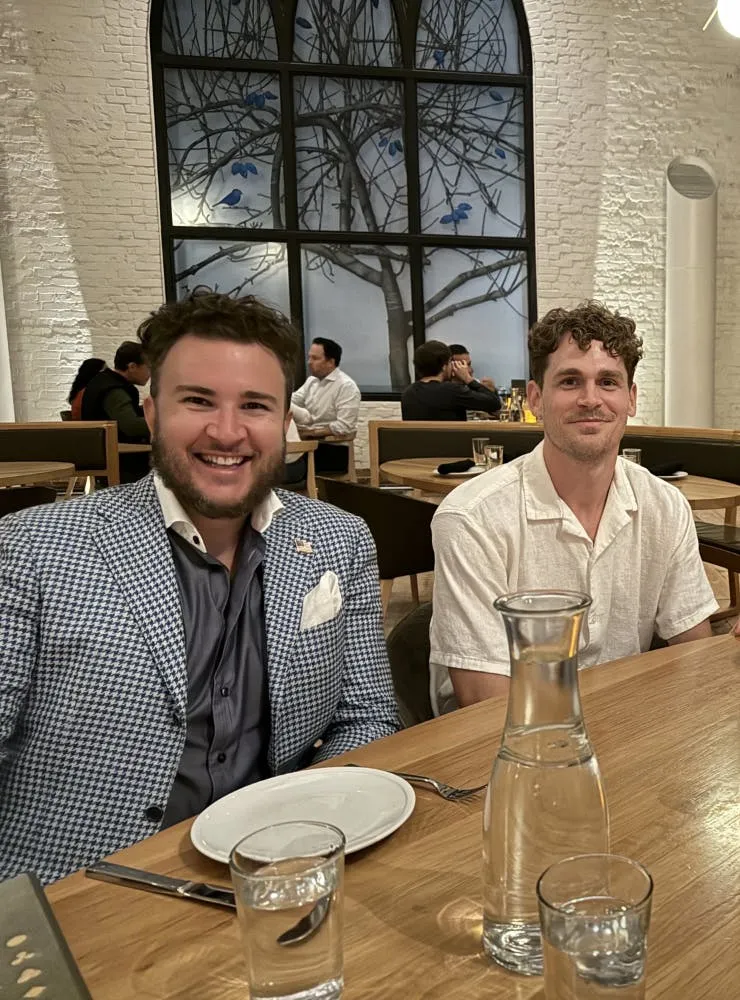 Two men smiling at a restaurant table with a water carafe and glasses in front of them, a large arched window with a bare-tree mural behind them