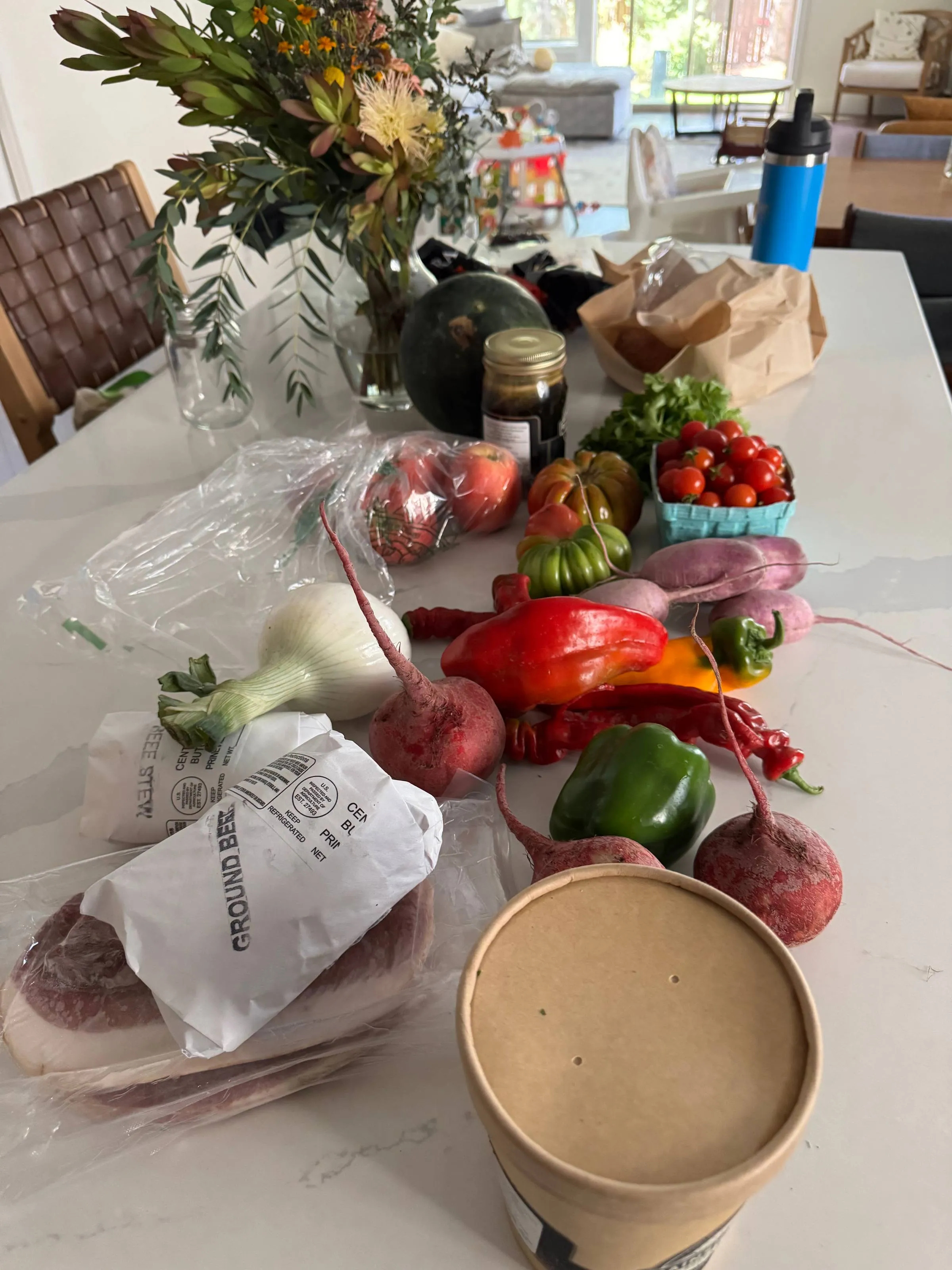 A kitchen counter covered with a farm haul: heirloom and cherry tomatoes, bell and chili peppers, radishes, beets, an onion, squash, greens, paper-wrapped ground beef and steak, a jar of preserves, and a bouquet of flowers