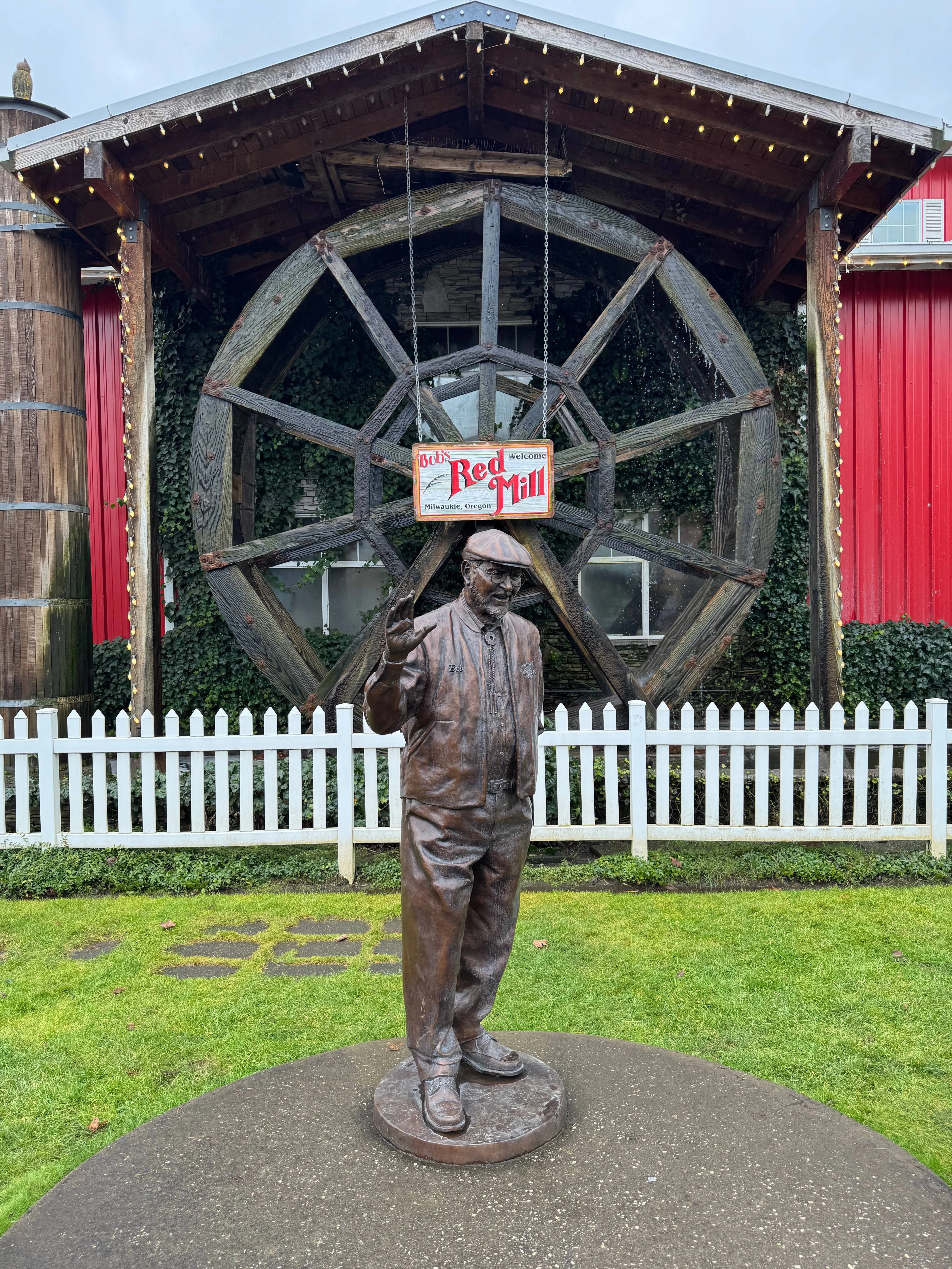 Bronze statue of Bob’s Red Mill founder Bob Moore waving, standing in front of a large wooden waterwheel with a “Bob’s Red Mill - Milwaukie, Oregon” welcome sign