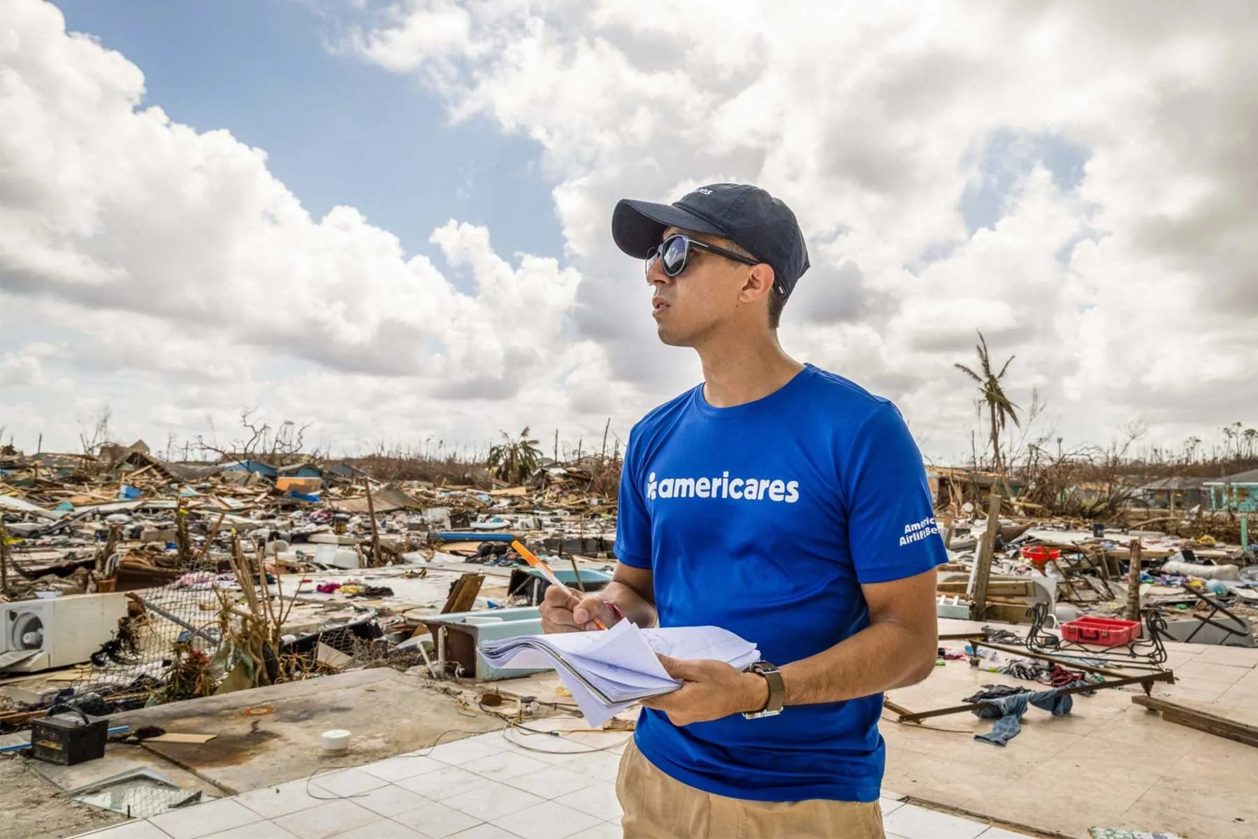 An Americares aid worker in a blue t-shirt and cap takes notes while standing amid the widespread rubble and debris of a disaster-struck area