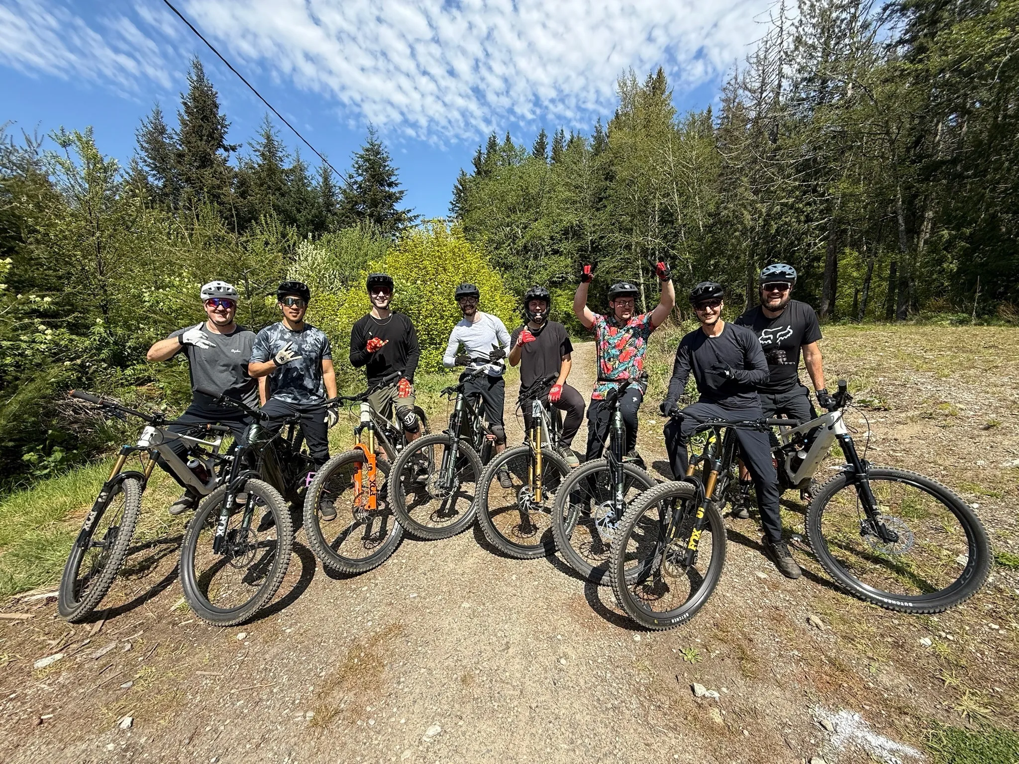 A group of eight mountain bikers posing with their bikes on a gravel trail surrounded by evergreen forest under a blue sky in Bellingham, Washington
