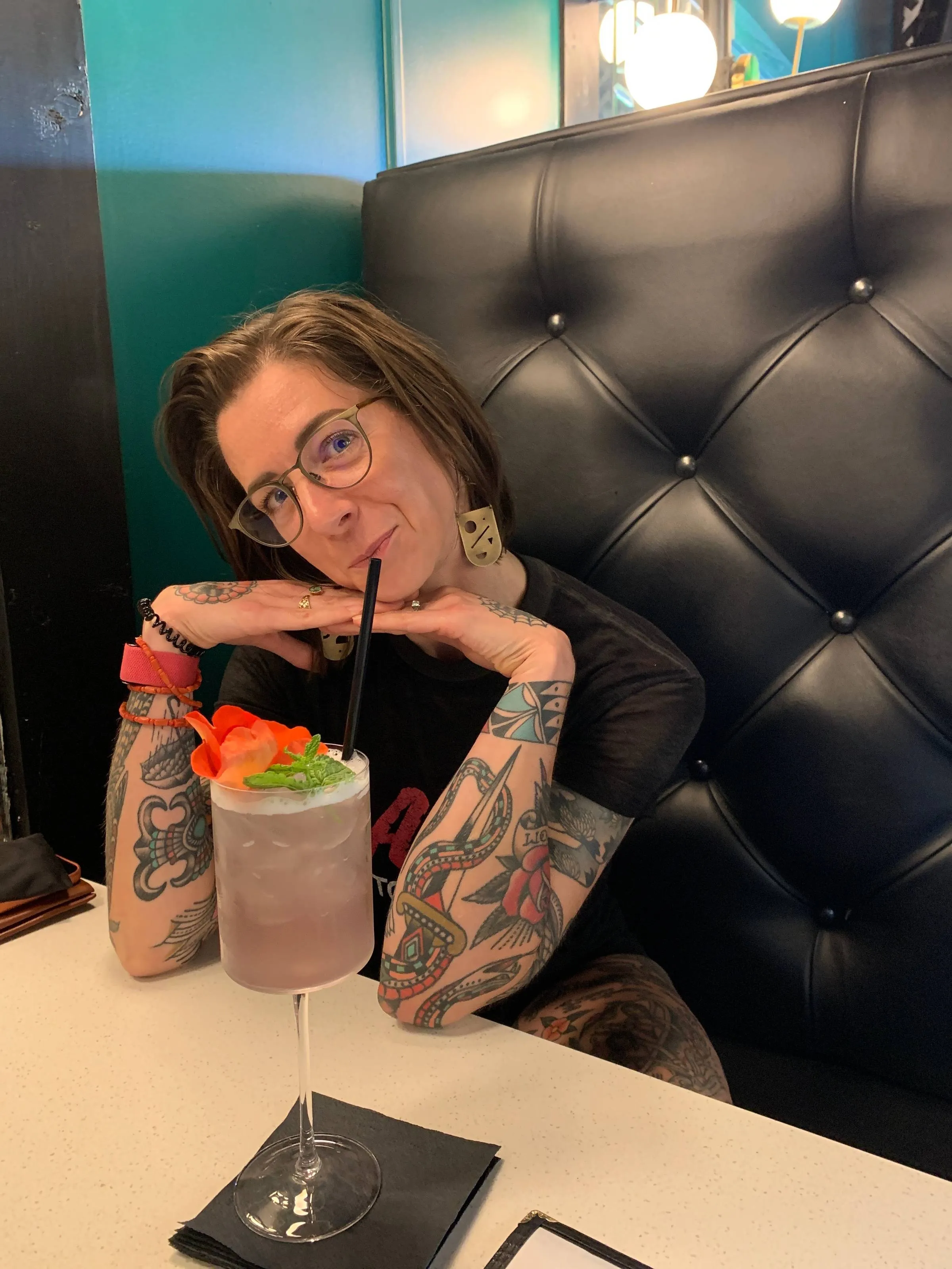 Sara, a woman with glasses and tattooed arms, resting her chin on her hands at a restaurant booth with a garnished cocktail in front of her