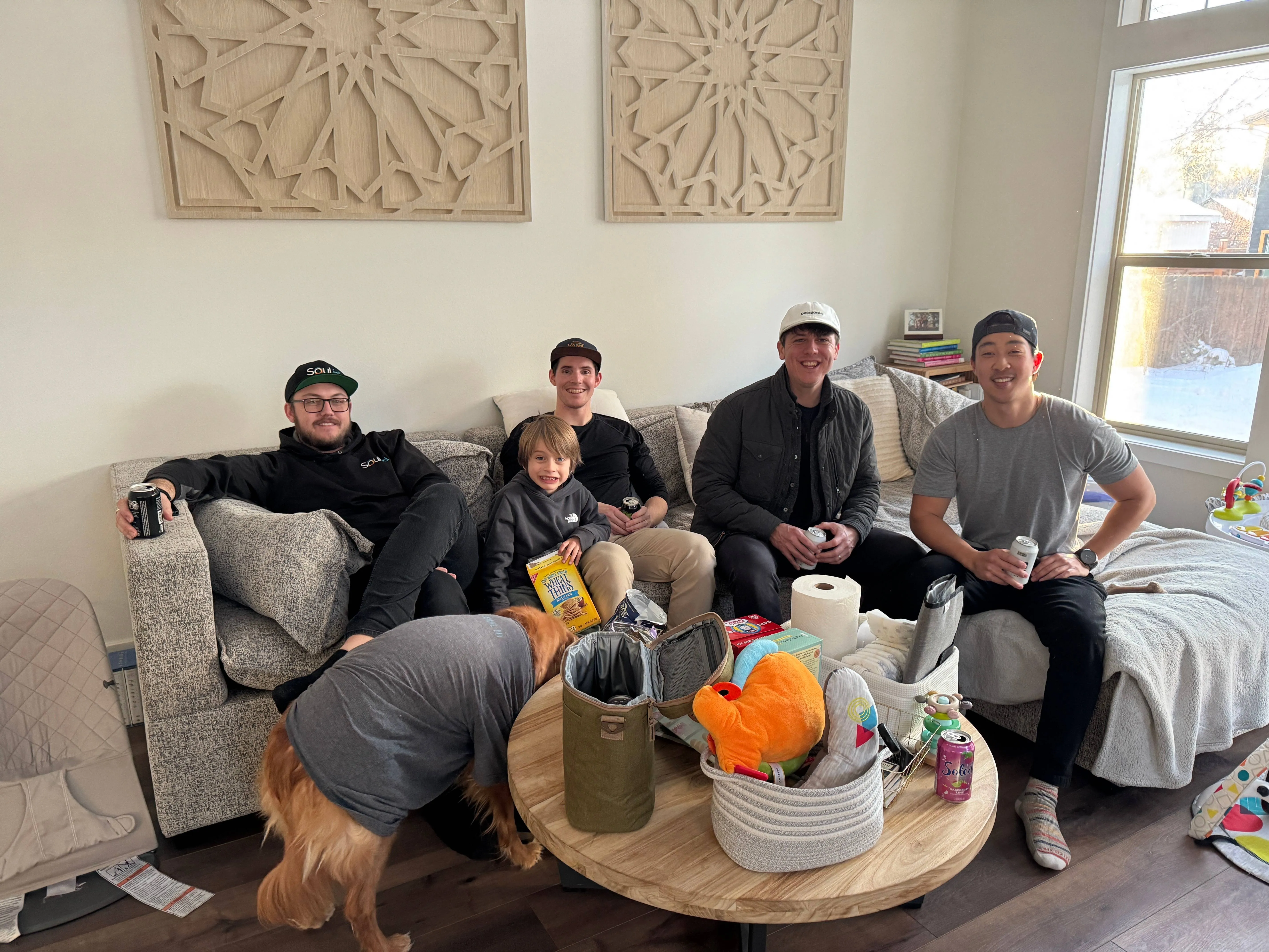 Four men and a young boy relaxed on a sectional couch with drinks and snacks spread across the coffee table while a golden retriever noses at the table
