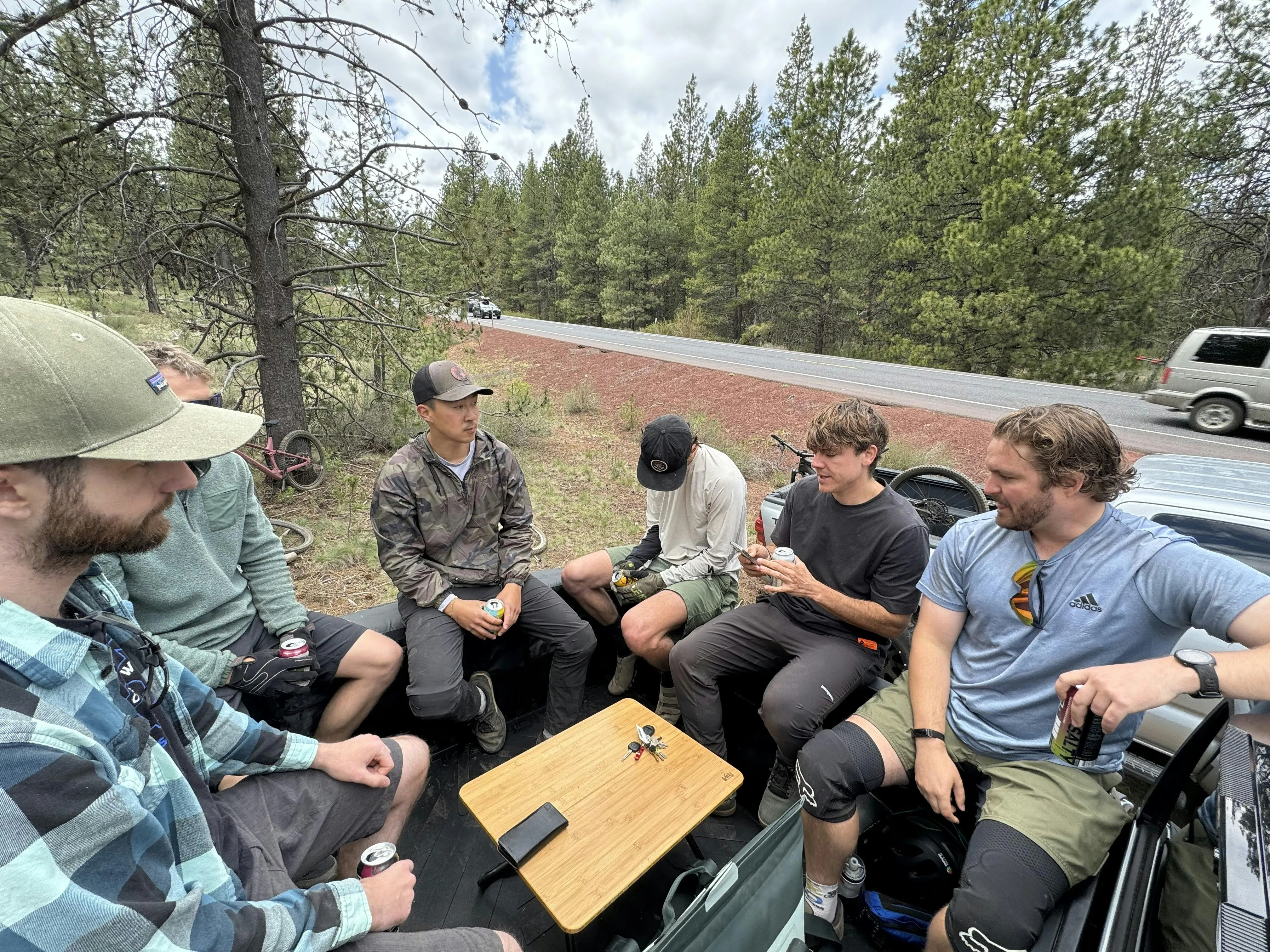 A group of mountain bikers sitting in a pickup truck bed drinking canned beers after a ride, with bikes leaning against pine trees along a rural road