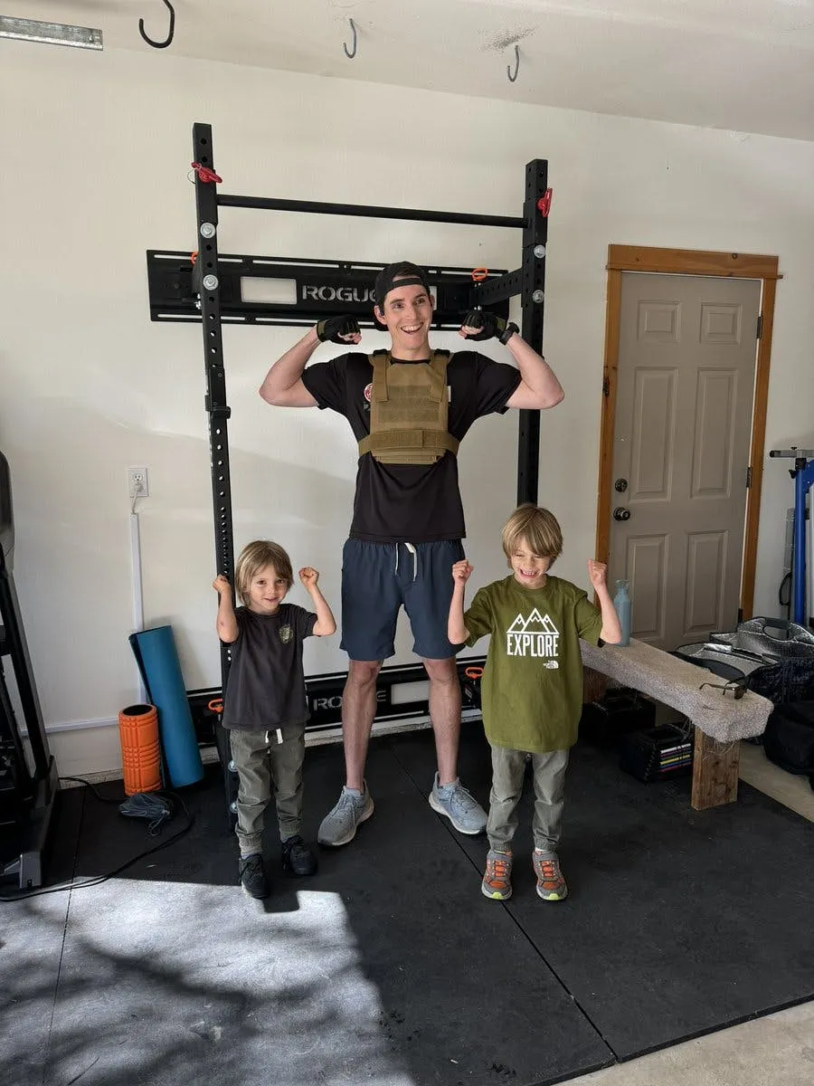 The author in a weighted vest flexing in a home garage gym after a workout, with two young kids flexing beside him