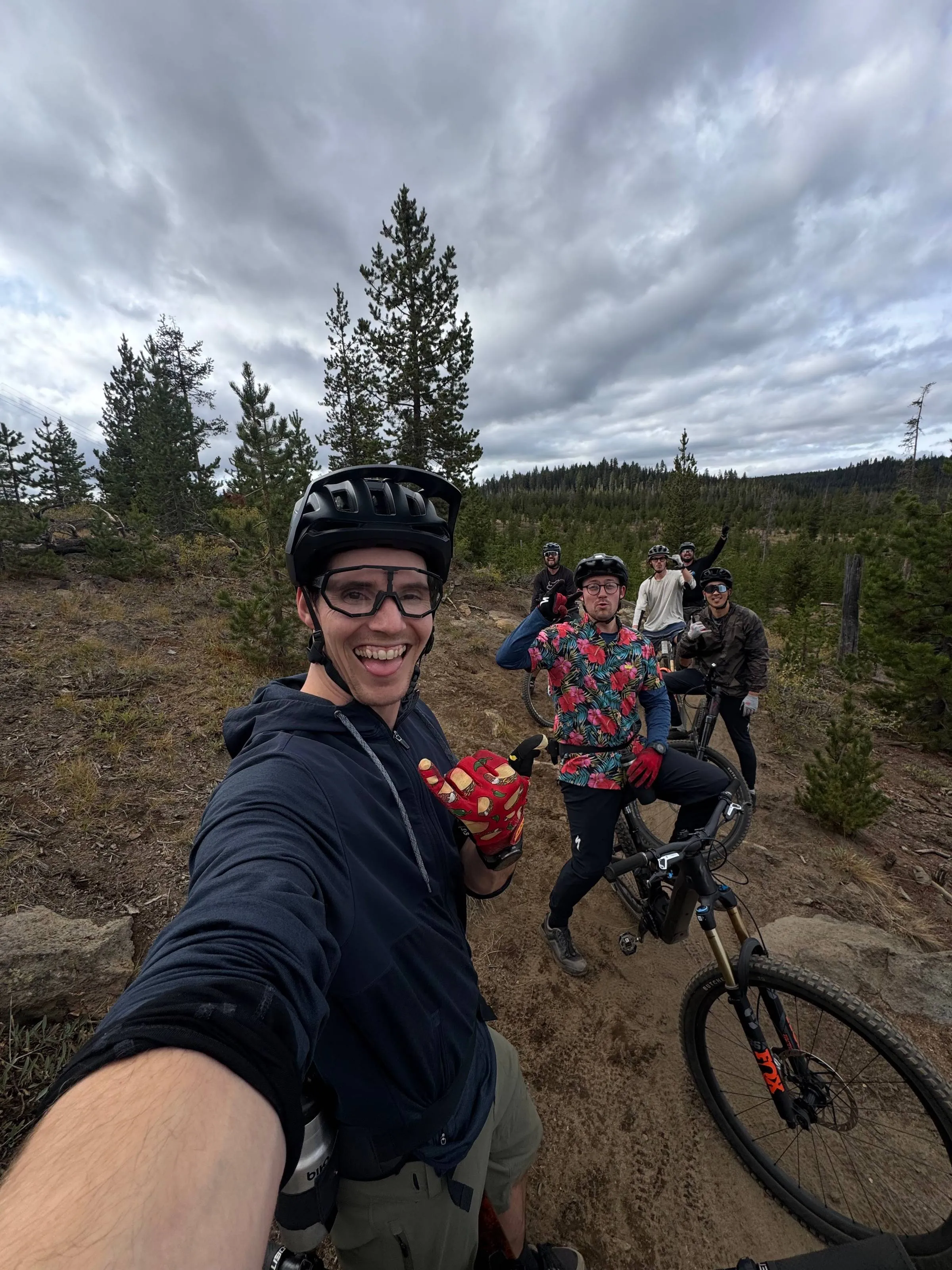 Selfie of a smiling man in a bike helmet on a mountain bike trail, with a group of friends on mountain bikes behind him among pine trees under a cloudy sky