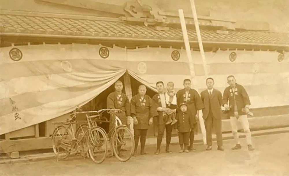 Sepia vintage photograph of a group of men and two children in traditional Japanese dress standing beside bicycles in front of a building draped with striped ceremonial fabric