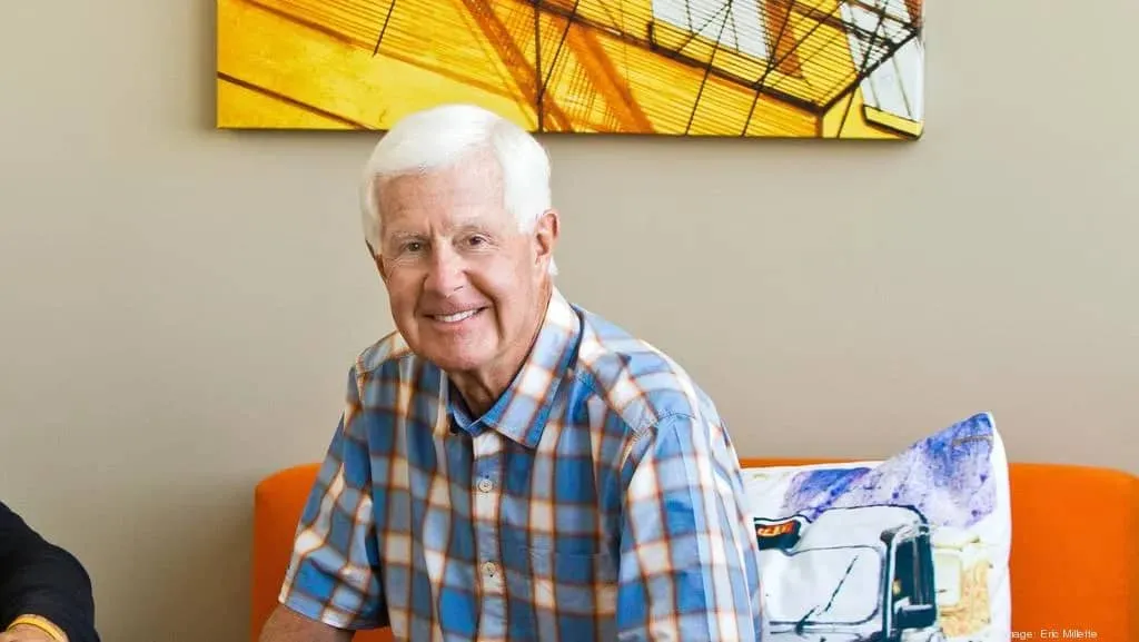 White-haired older man in a blue plaid shirt smiling while seated on an orange couch beneath a yellow abstract painting