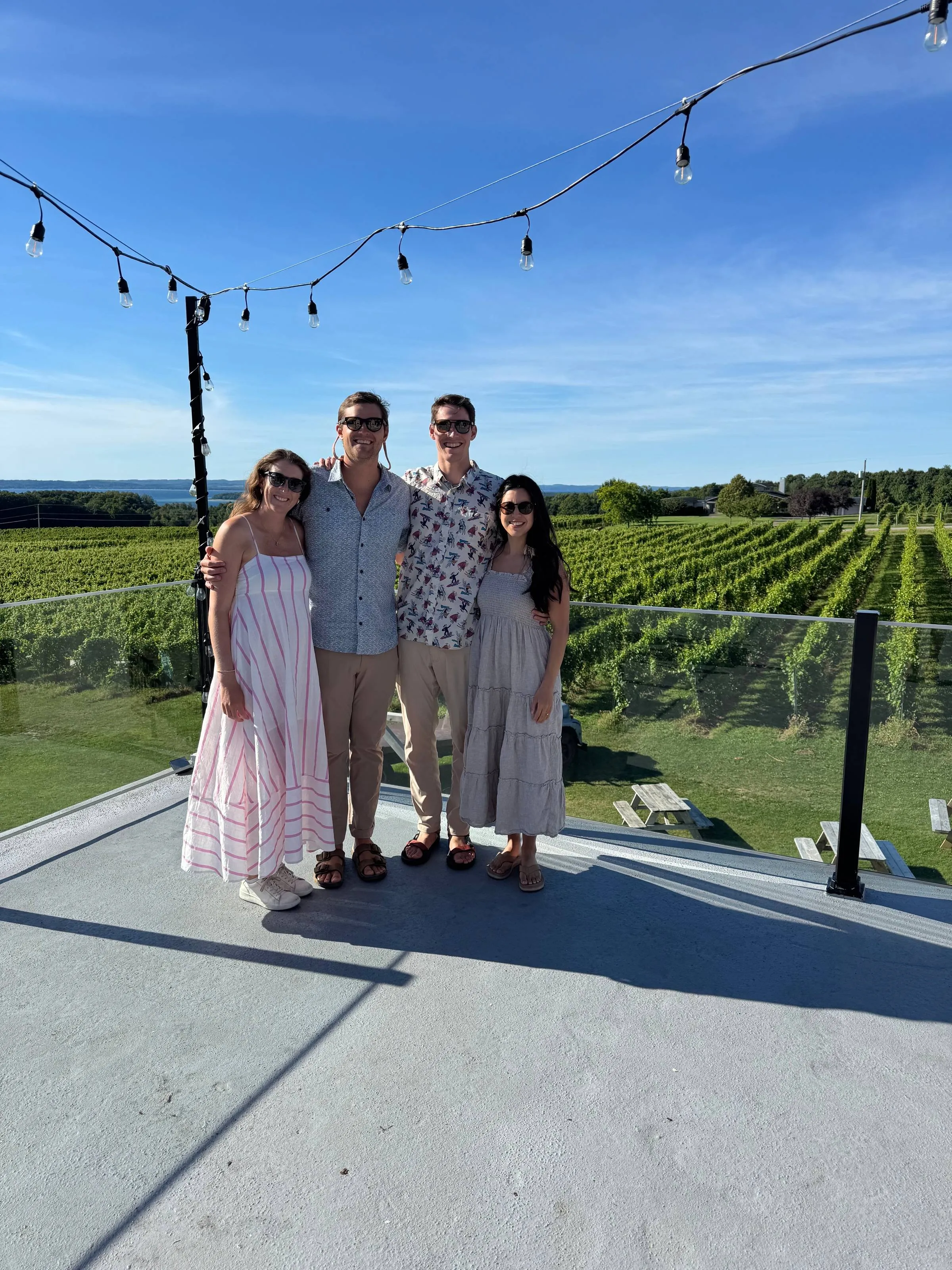 Two couples posing arm in arm on a deck overlooking a sunlit vineyard and lake in Traverse City, with string lights hanging above