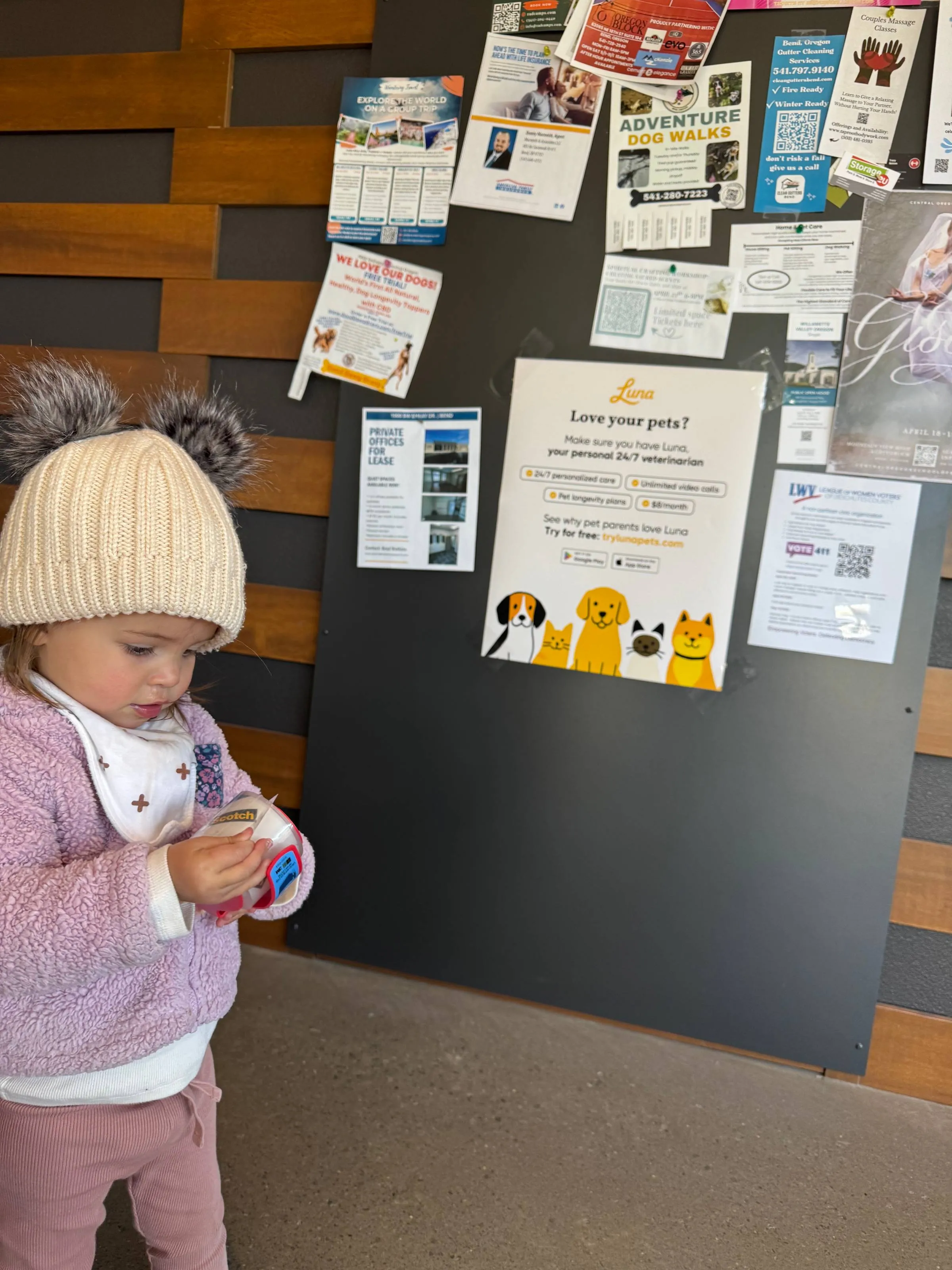 Toddler in a knit pom-pom hat holding a tape dispenser beside a community bulletin board featuring a Luna pet-care poster