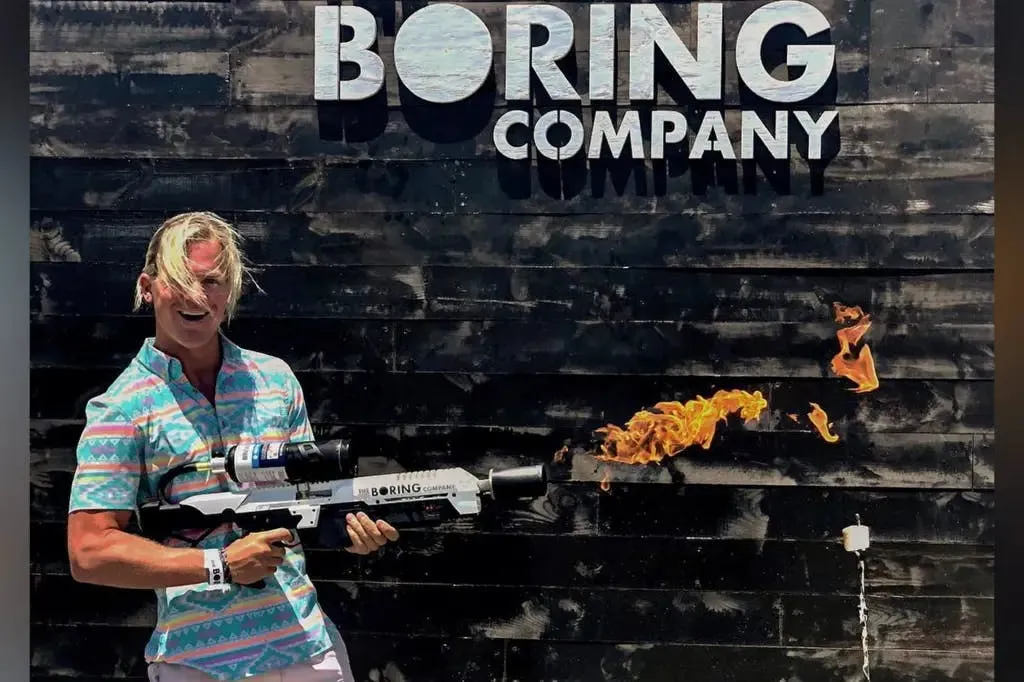 A grinning person firing The Boring Company’s branded flamethrower in front of a black wall reading “Boring Company”