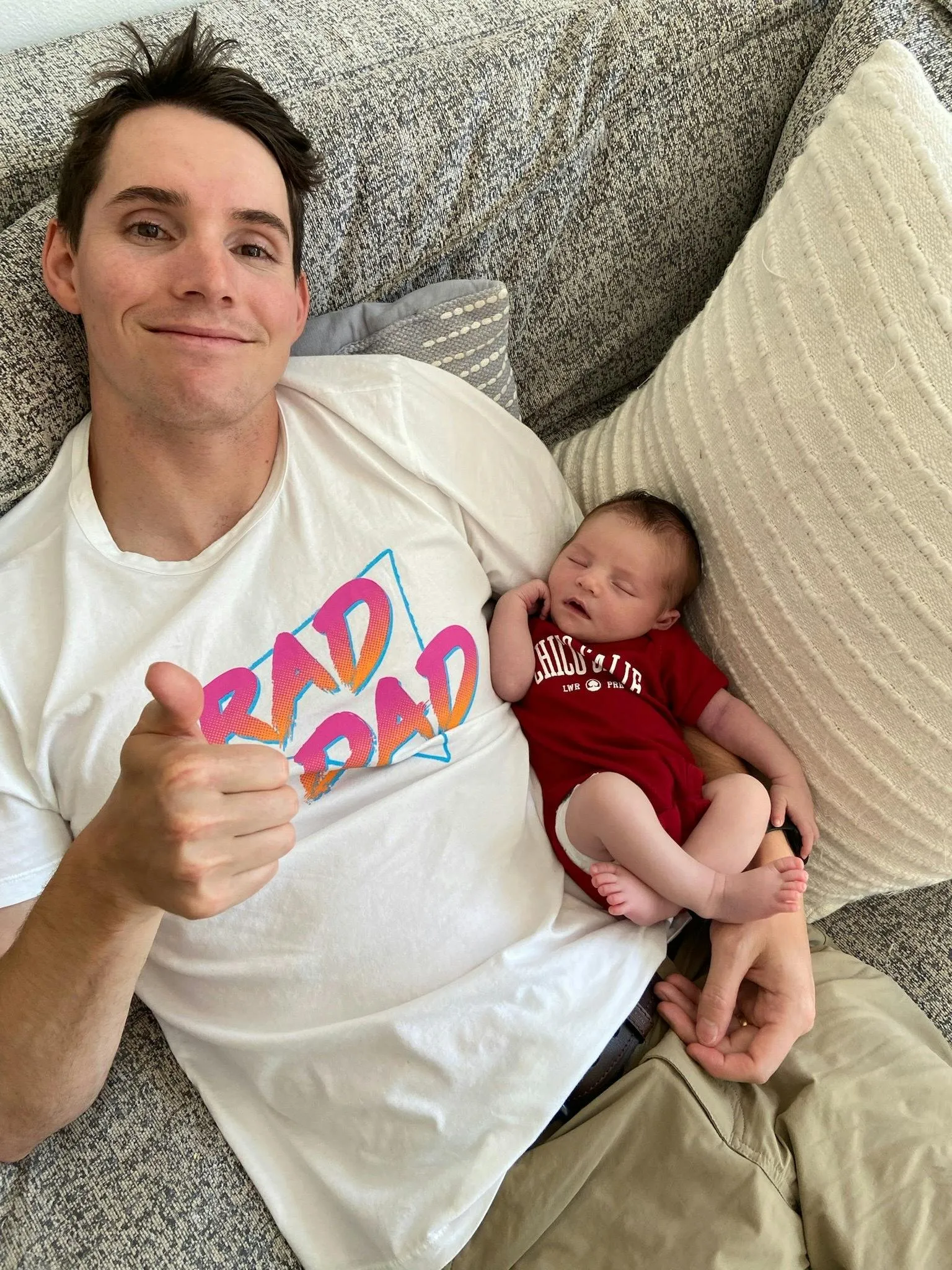 Ben lying on a couch giving a thumbs up in a white βRad Dadβ t-shirt with a sleeping newborn baby in a red onesie resting on his arm