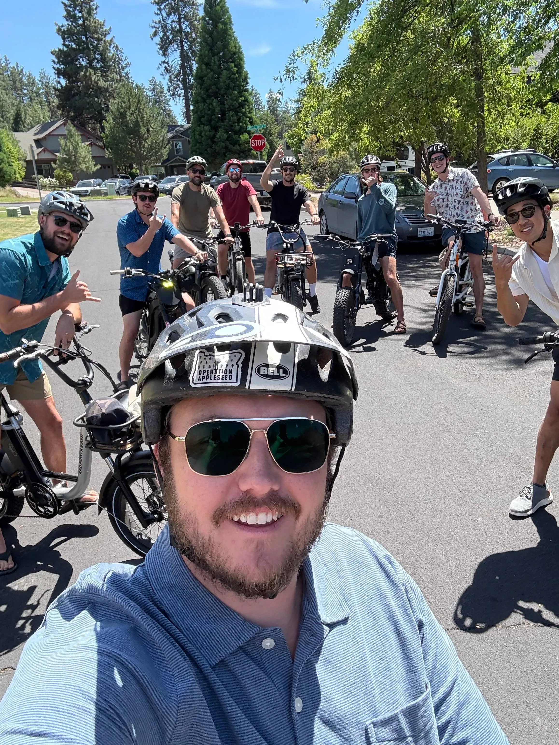Group selfie of nine men in bike helmets on e-bikes on a sunny suburban Bend street, several throwing shaka and rock-on hand signs