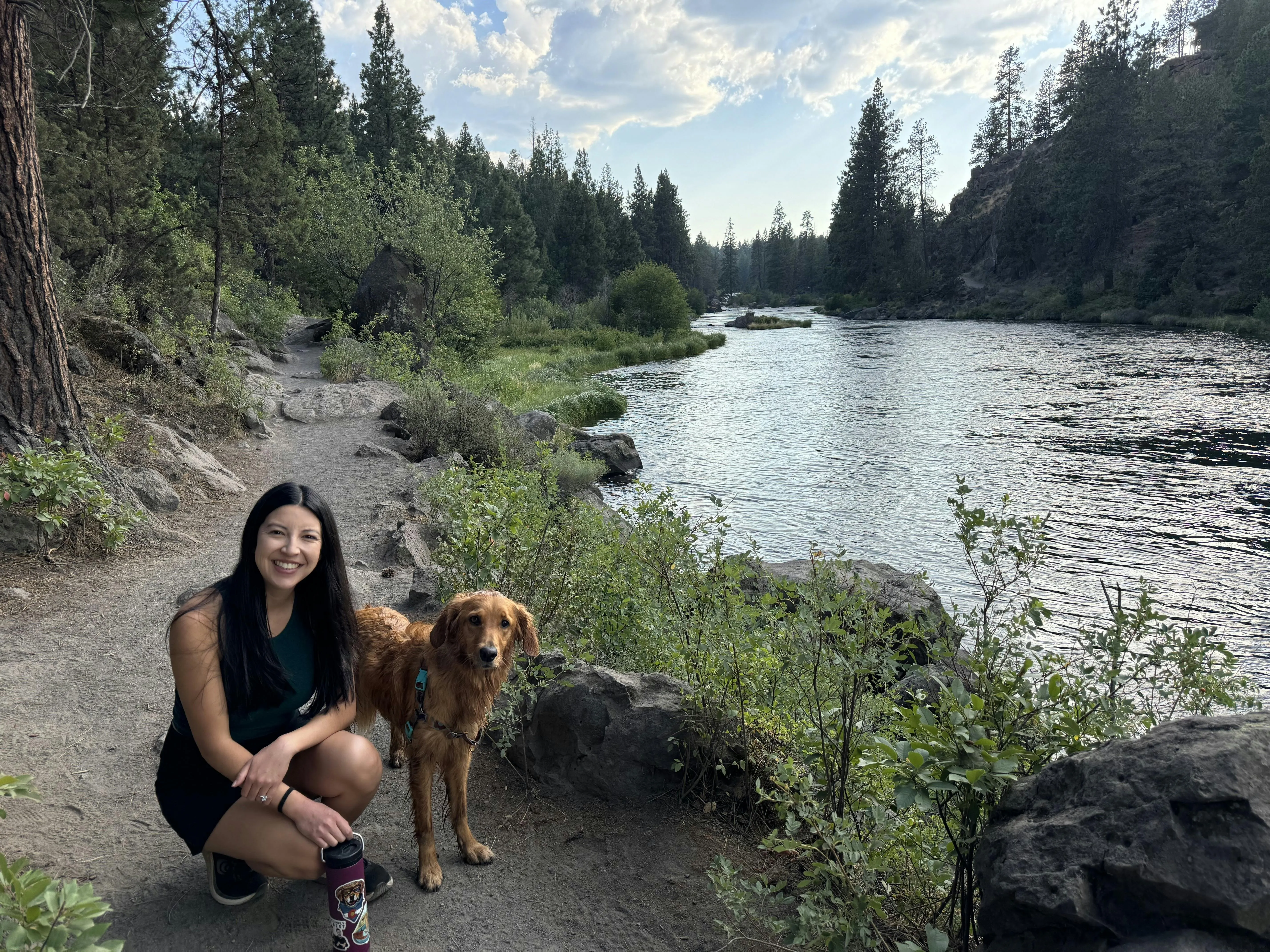A woman crouching beside a wet golden retriever on a riverside trail lined with pine trees in evening light