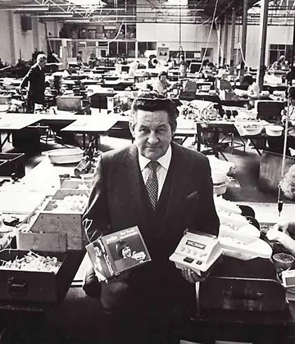 Black-and-white photo of Thomas Cassidy in a suit on the Casdon factory floor, holding two boxed toys, with rows of workers assembling products behind him
