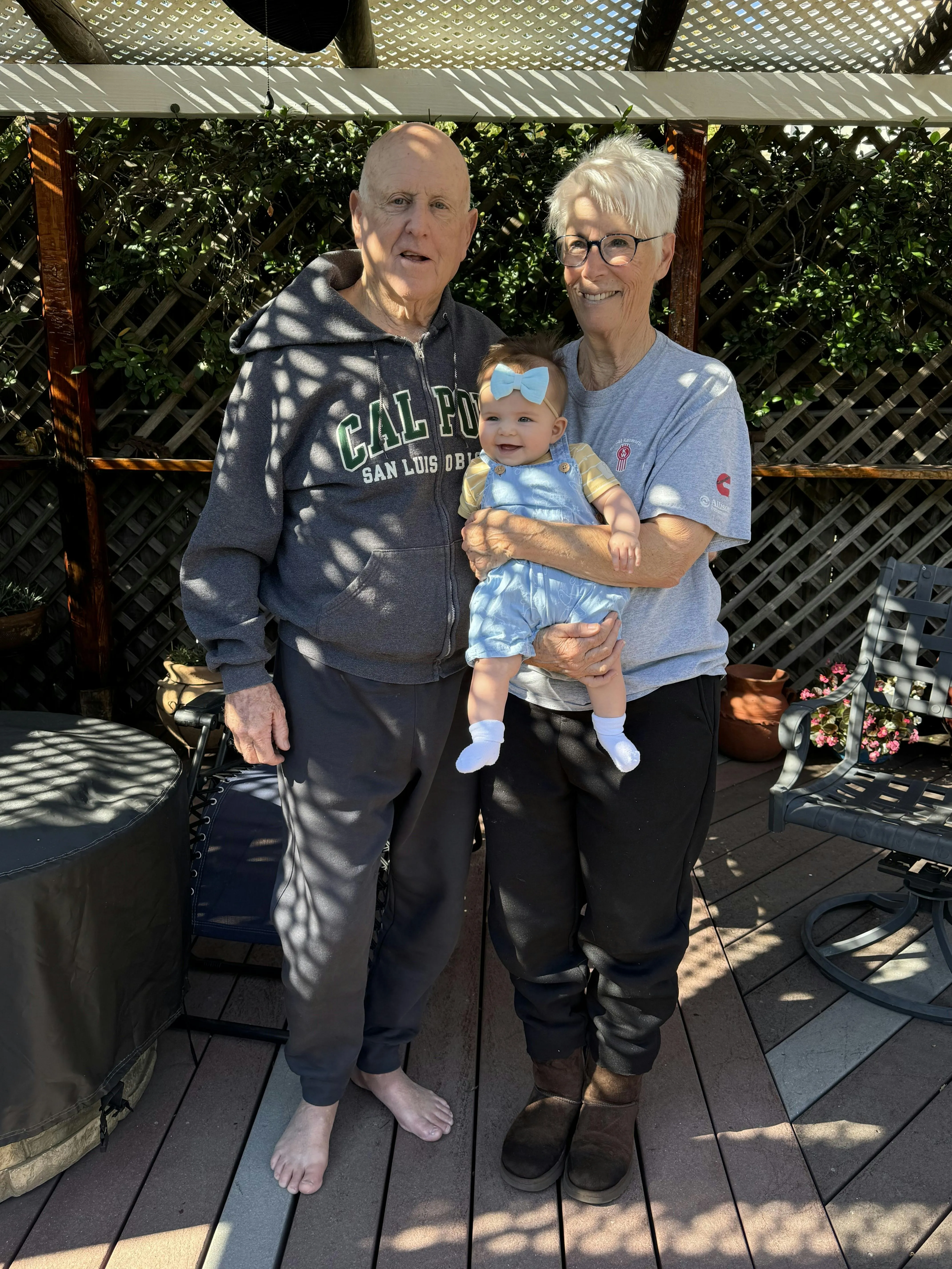 Great-grandparents standing on a sunny backyard deck, the grandmother holding a smiling baby wearing a light blue bow and overalls