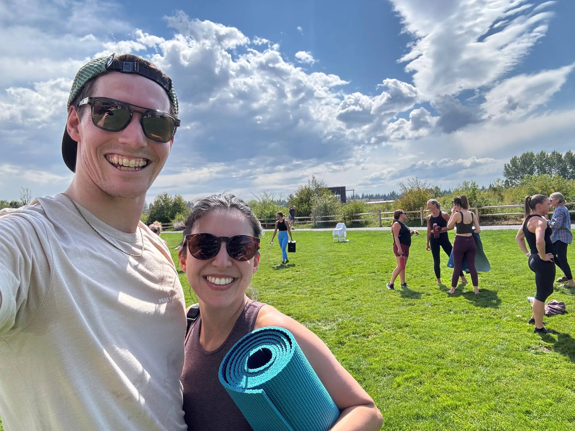 Outdoor selfie of Ben and Amelia in sunglasses, Amelia holding a rolled blue yoga mat, with a group of people gathered on a grass field for a barre class behind them