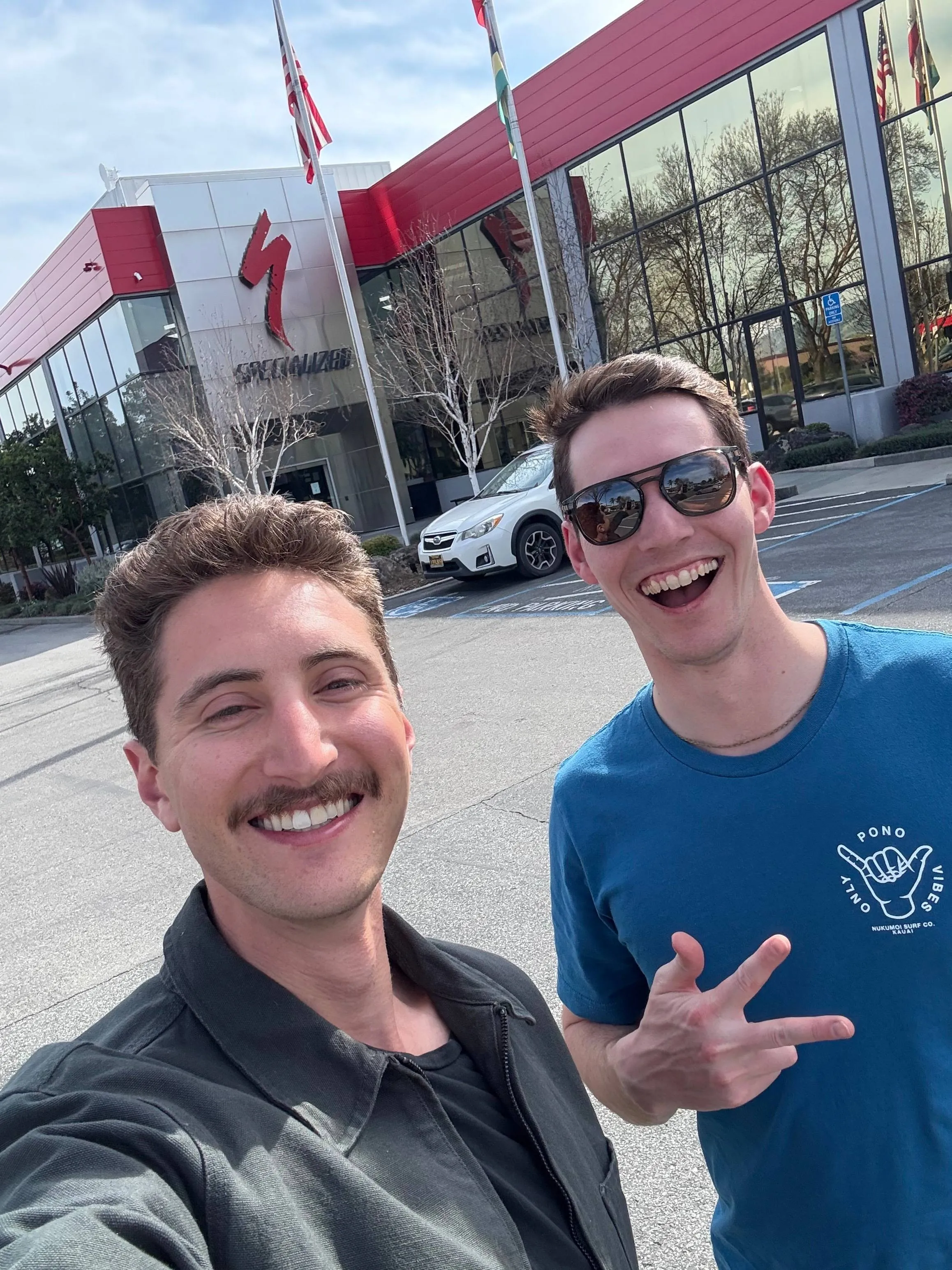 Two smiling men taking a selfie in the parking lot outside the Specialized Bikes headquarters, with the red Specialized “S” logo on the building behind them