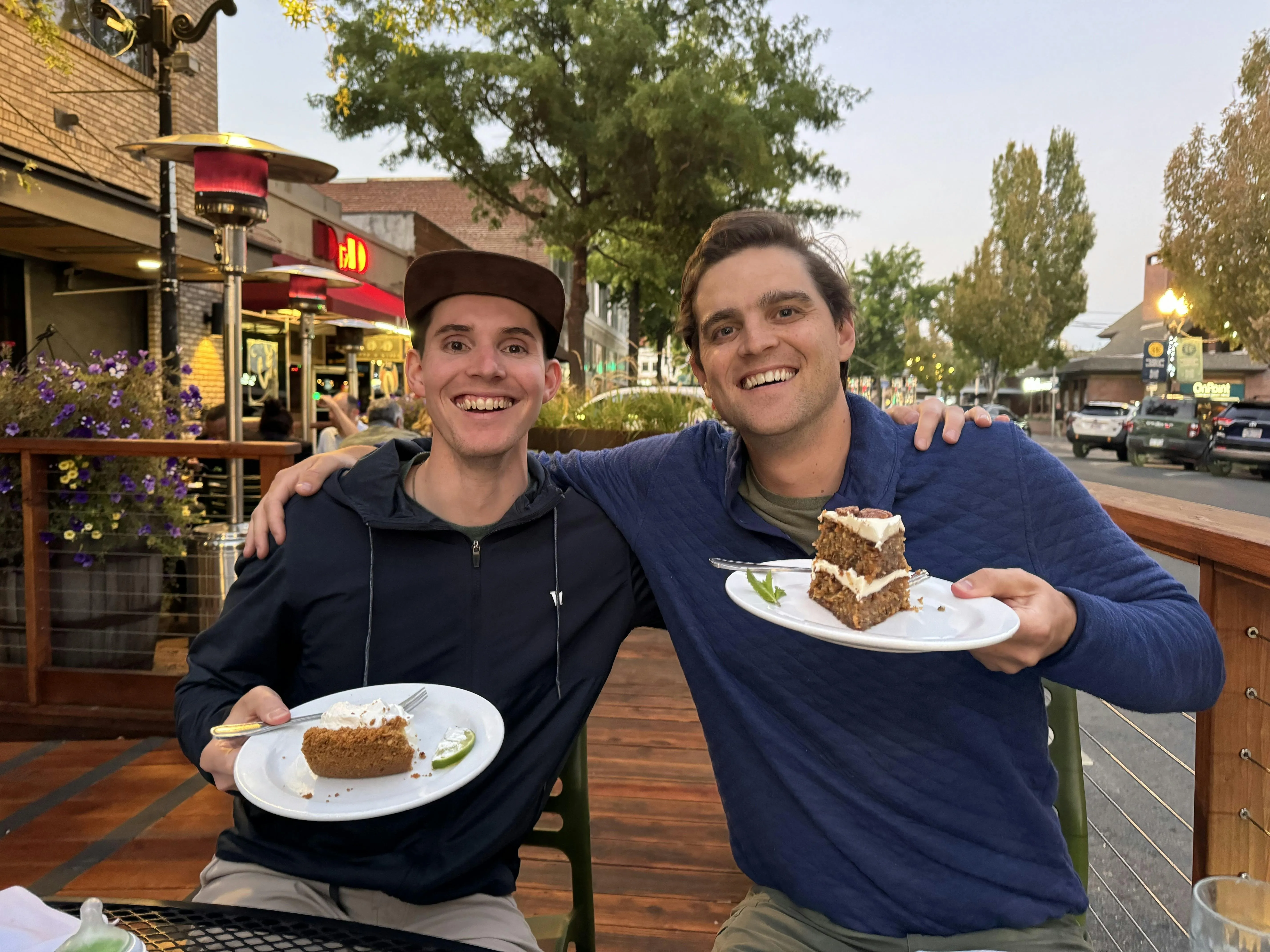 Ben and his business partner Andy smiling side by side at an outdoor restaurant table at dusk, each holding a plate with a slice of cake