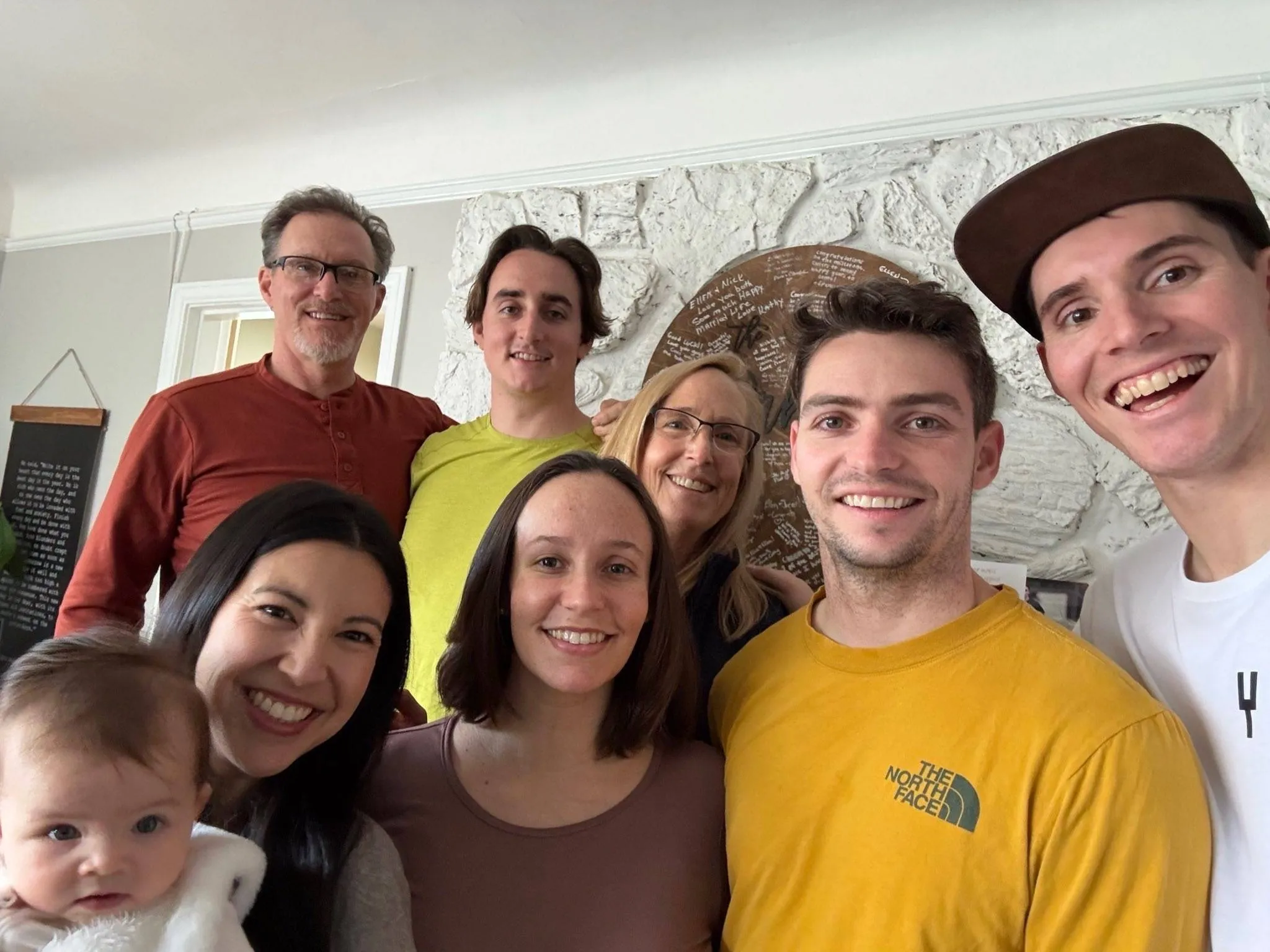 Group selfie of a smiling family of eight, including a baby, gathered indoors in front of a white stone fireplace