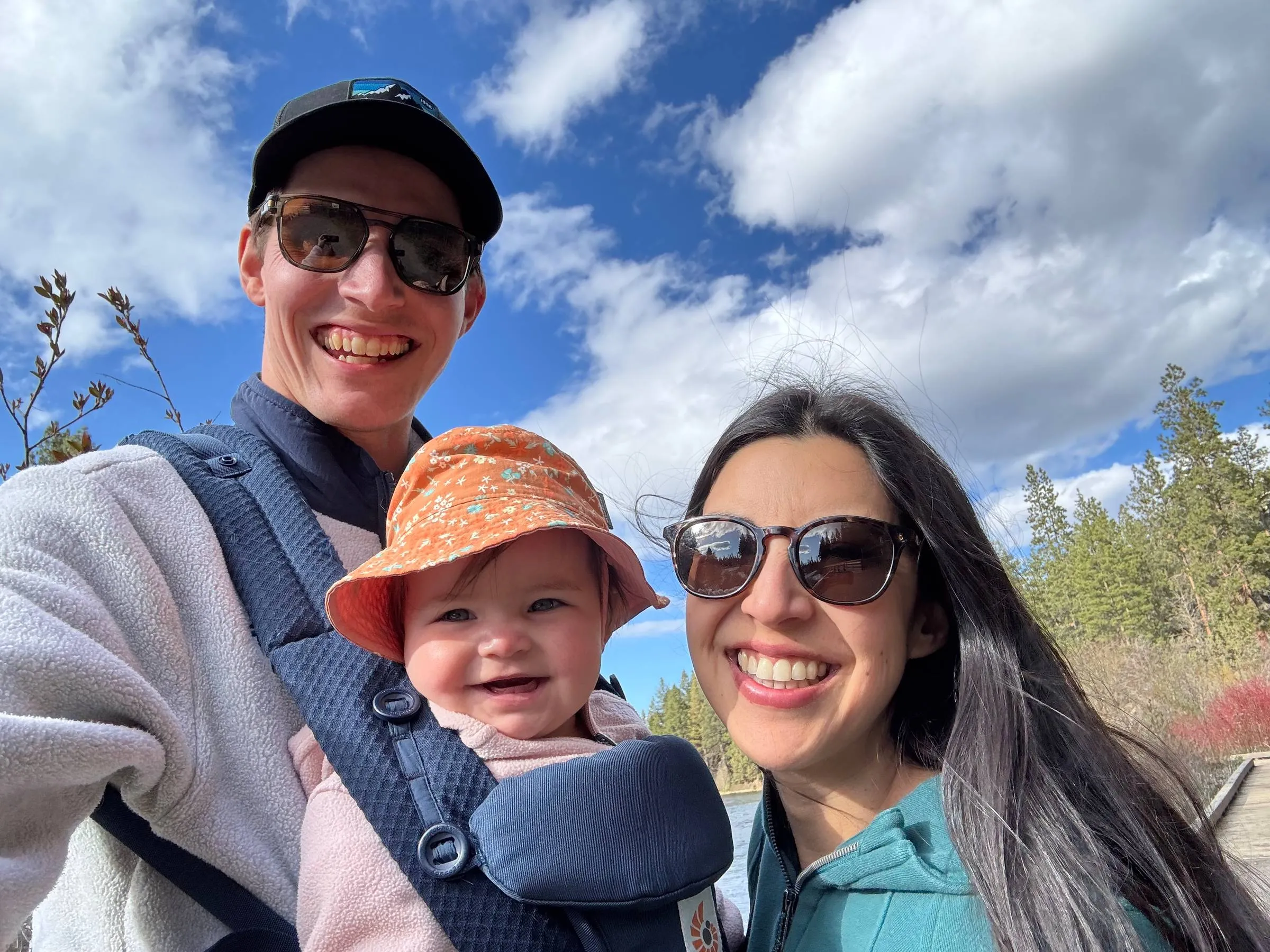 Ben taking a selfie outdoors by a lake with his partner and their smiling baby in a front carrier, on a sunny day with blue skies and pine trees