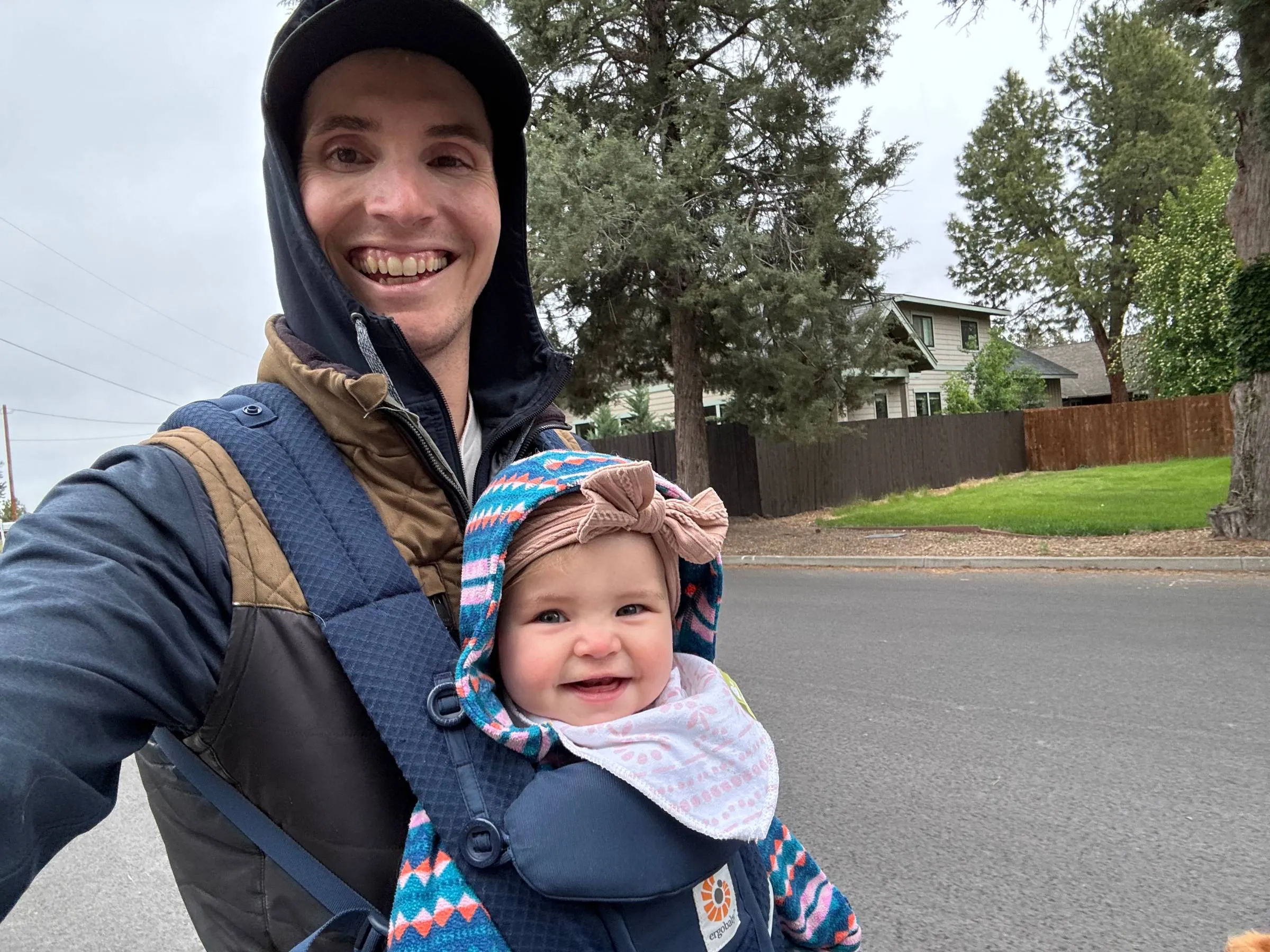 Smiling man in a hooded jacket taking a selfie on a residential street with a happy baby wearing a bow headband strapped to his chest in a carrier