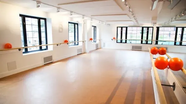 Empty barre studio with a light wood floor, wall-mounted ballet barres, and orange exercise balls