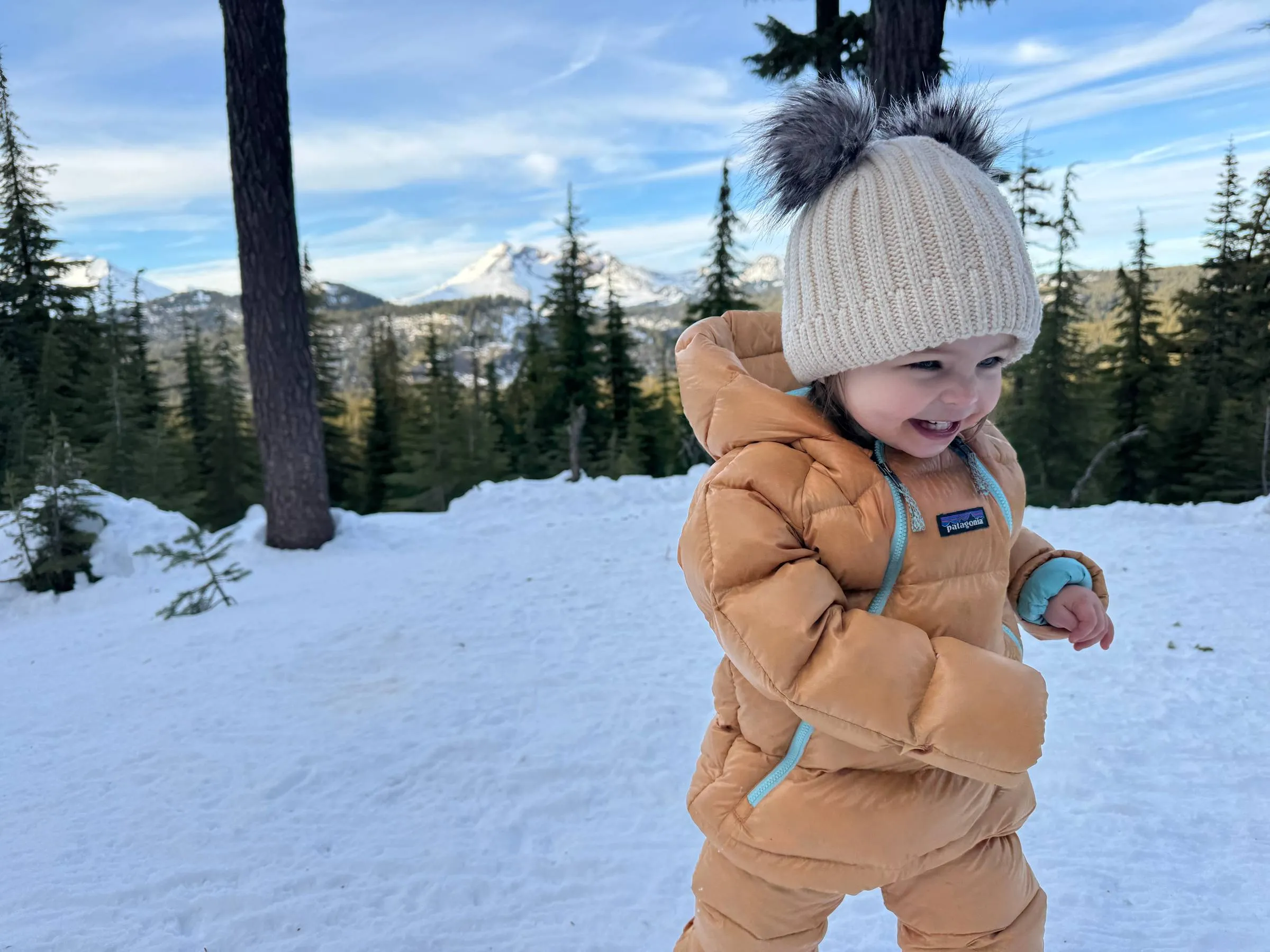 Toddler in a tan snowsuit and pom beanie smiling in the snow with pine trees and mountains behind