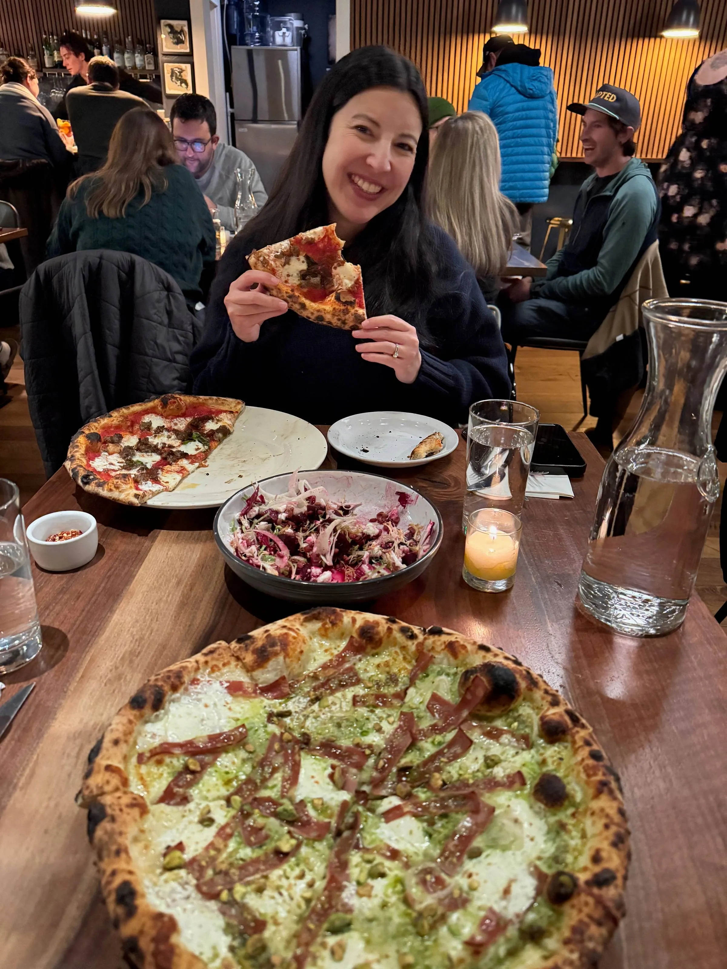 Woman smiling and holding a slice of pizza at a restaurant table full of pizzas and a salad