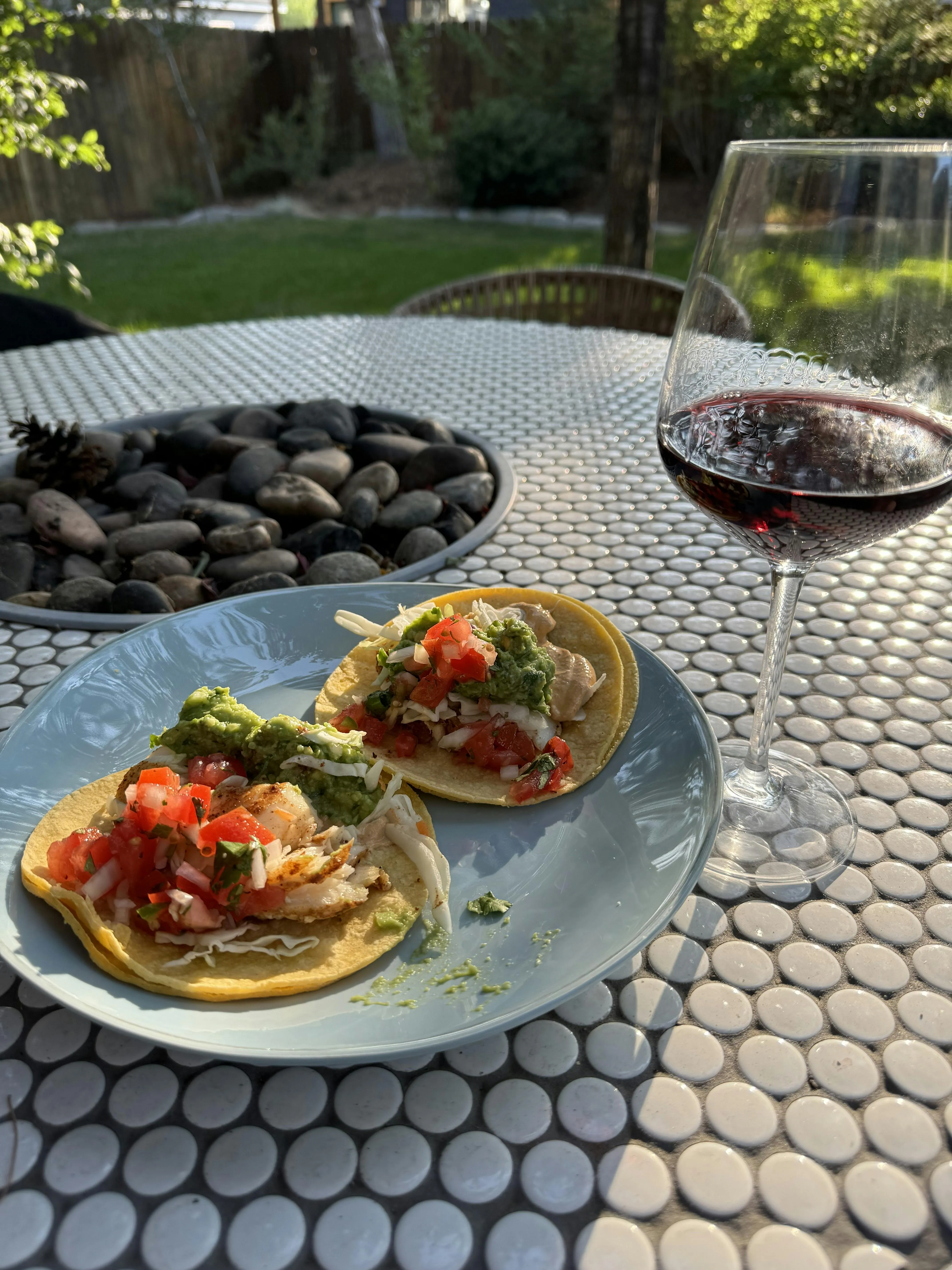 Two homemade fish tacos topped with pico de gallo and guacamole on a plate, next to a glass of red wine on a patio table