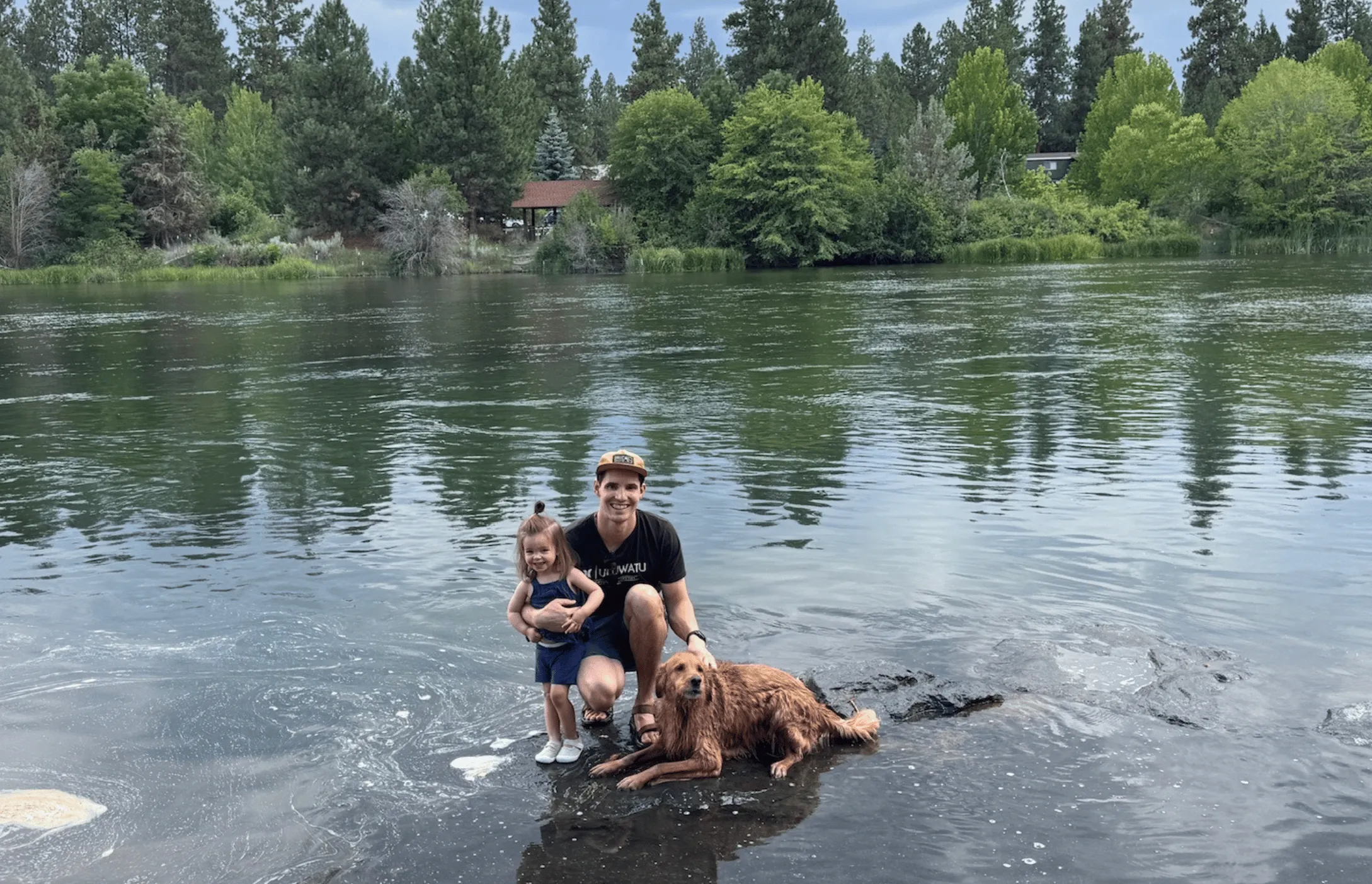 Ben crouching on a rock in a river with his toddler daughter and a wet golden retriever, pine trees along the far bank