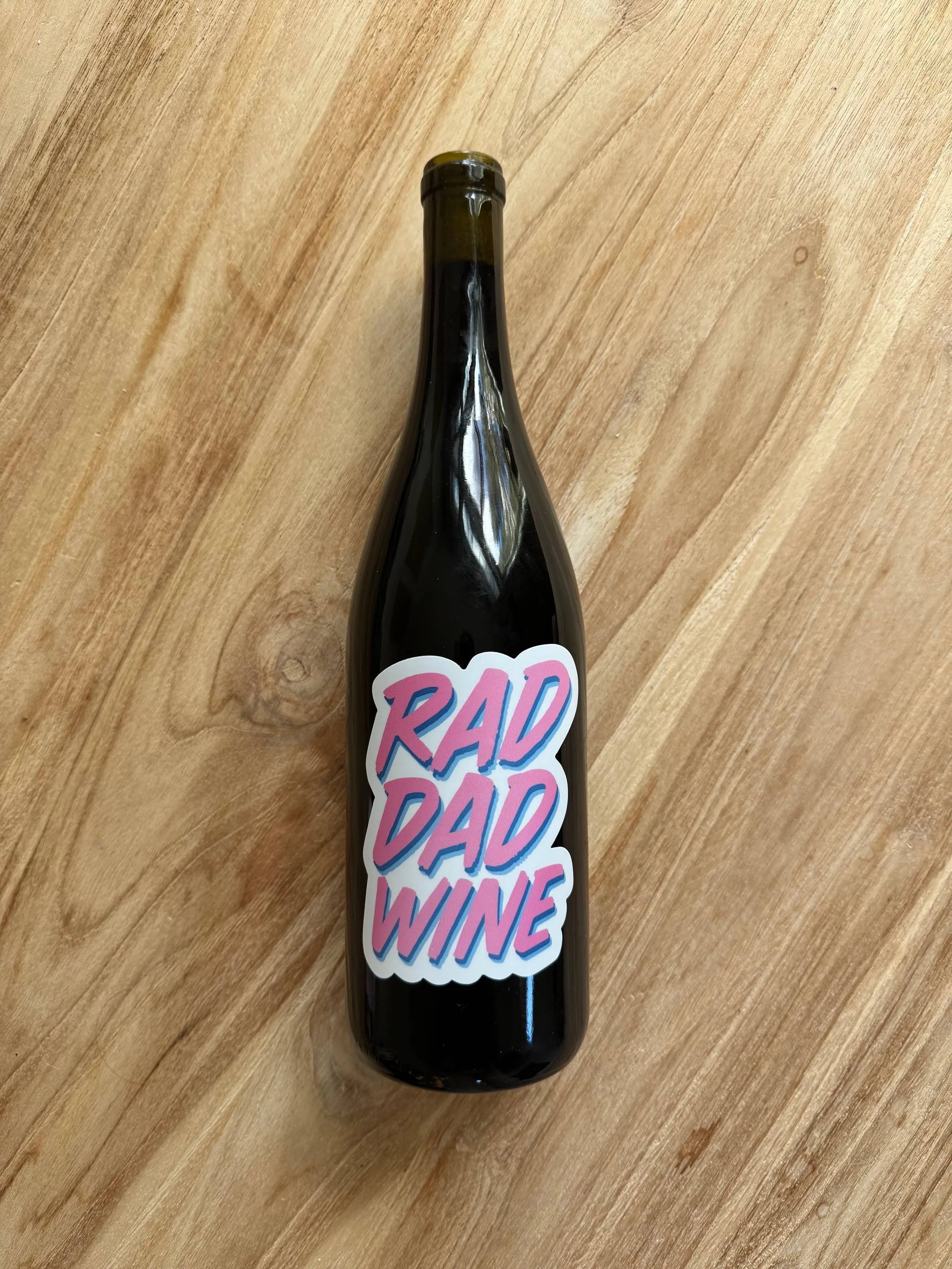 A dark bottle of wine lying on a wooden surface with a pink “RAD DAD WINE” sticker label