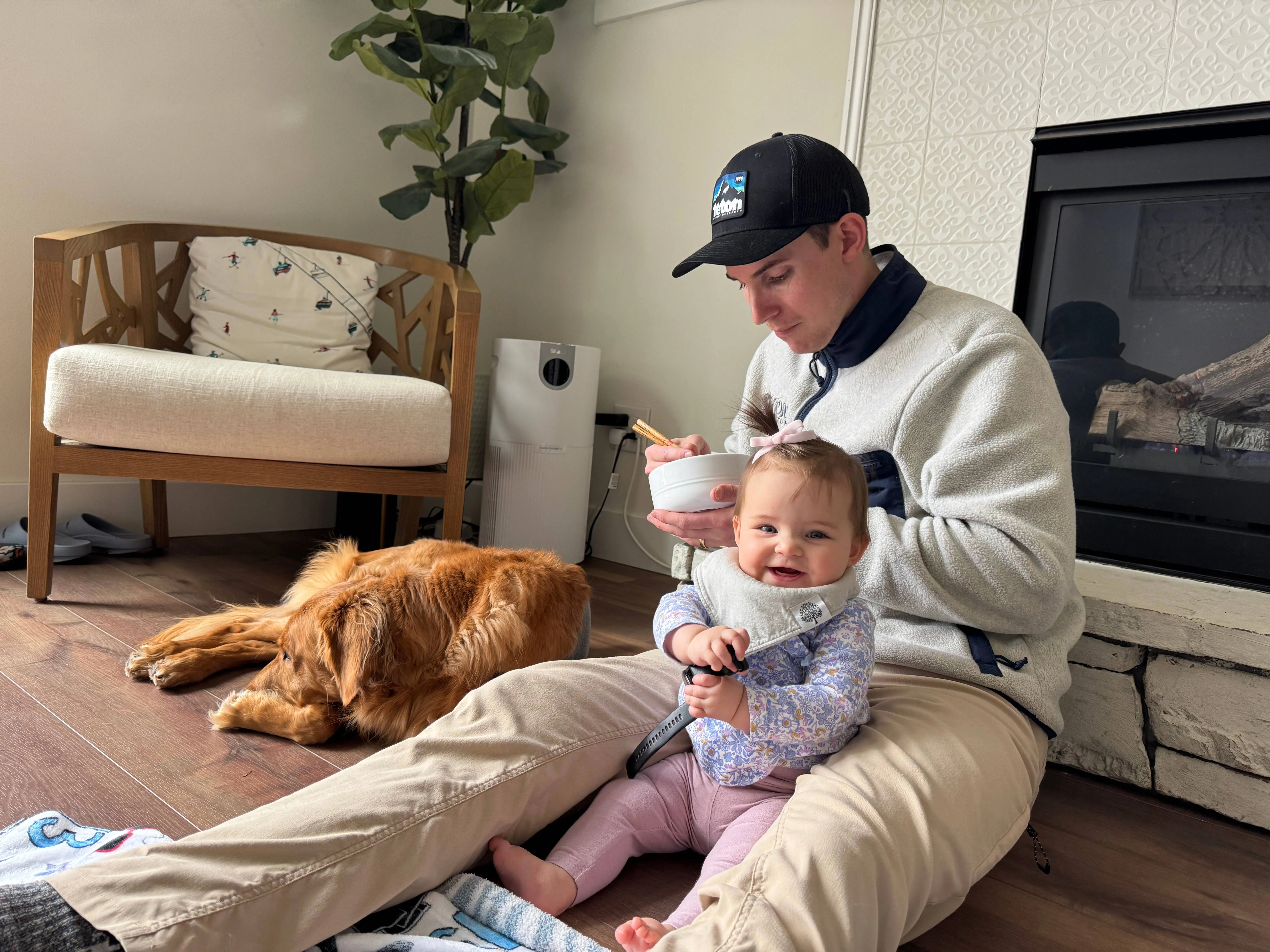 Man in a cap and white fleece feeding a smiling baby girl from a bowl while a golden retriever lies on the living room floor beside a fireplace