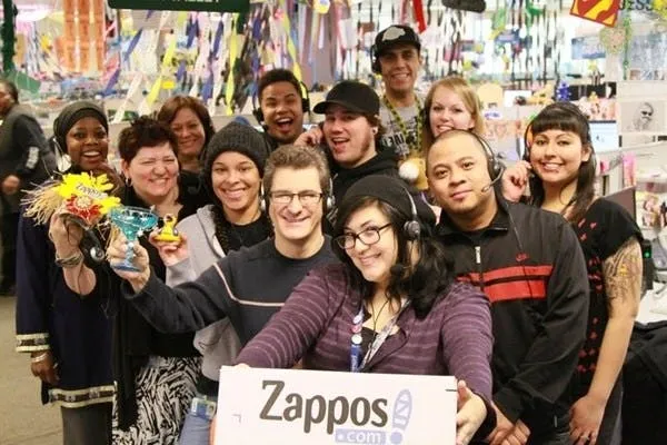 A group of Zappos employees crowding together for a smiling selfie, one holding up a Zappos.com sign