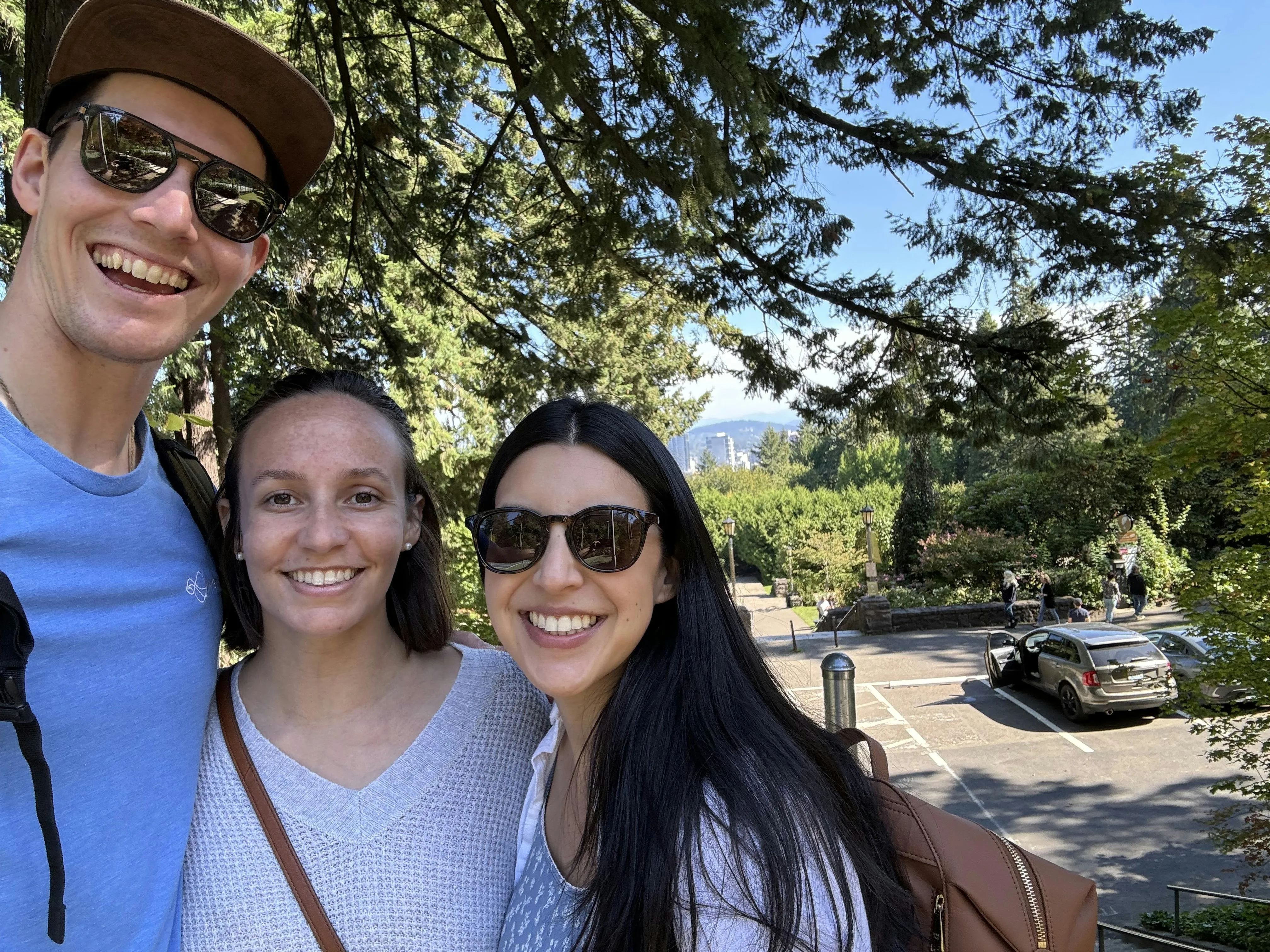 Selfie of Ben with two women under evergreen trees at a Portland park, with the city skyline visible in the background