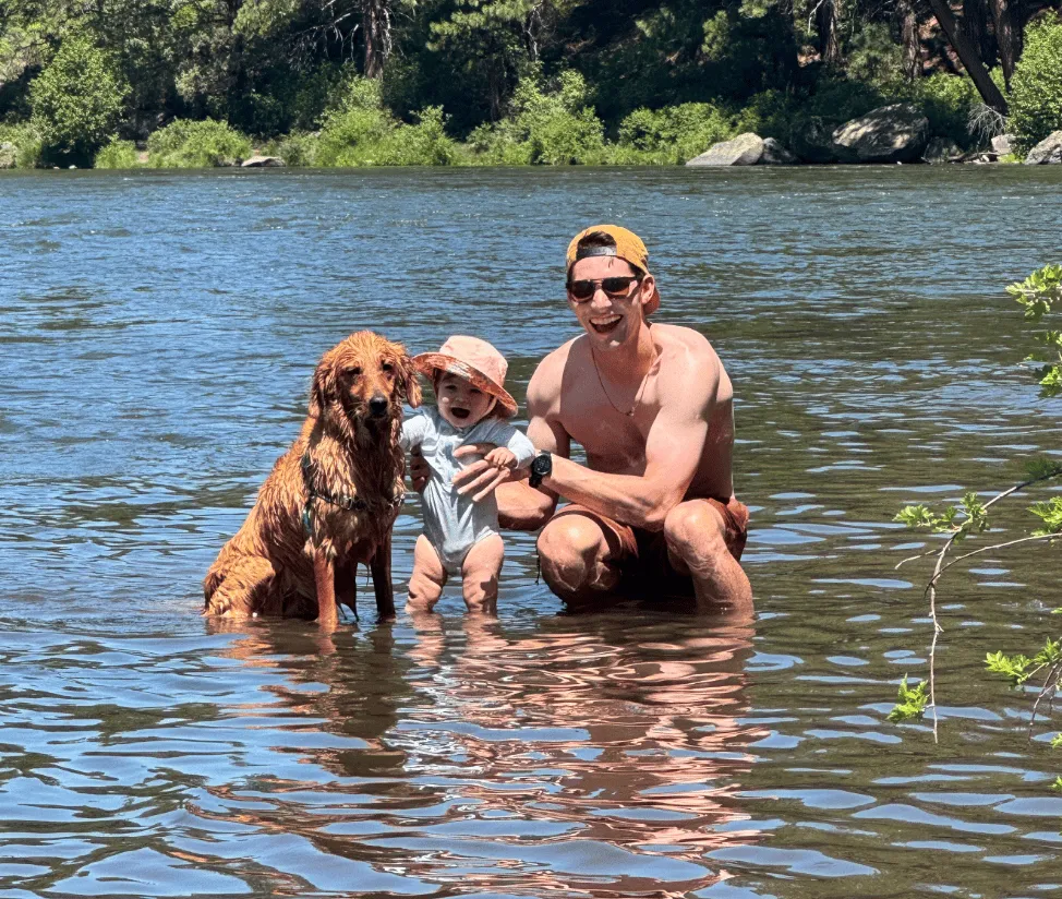 Ben crouching in a river with a smiling baby in a sun hat and a wet golden retriever