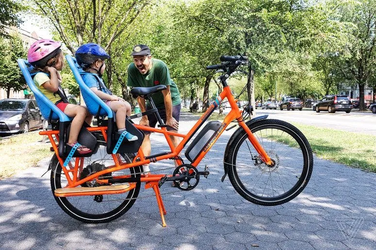 A man grinning at two helmeted kids seated on the back of an orange Rad Power electric cargo bike on a tree-lined path