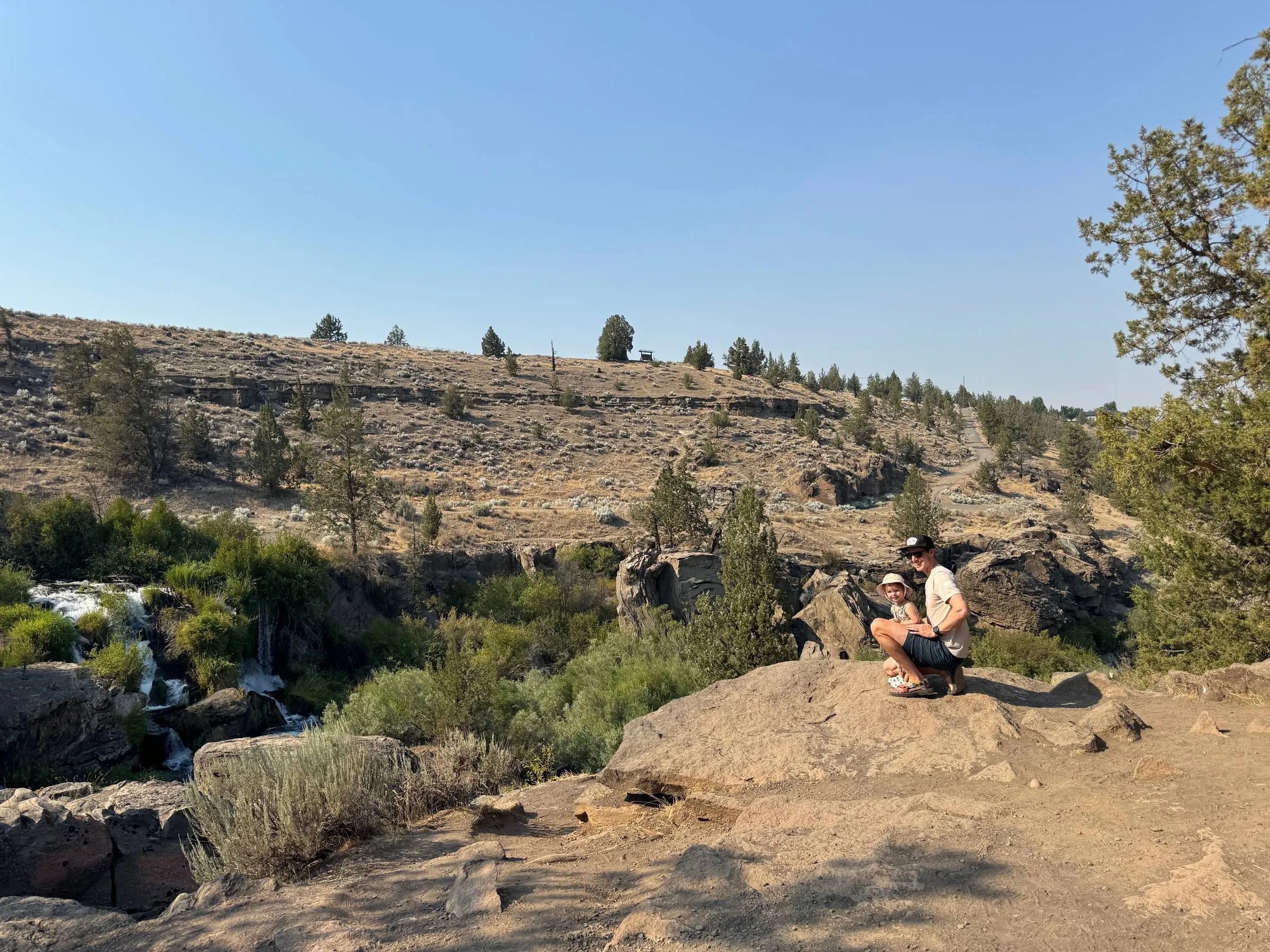 Ben crouching with his toddler daughter on a rock ledge above a desert waterfall, with a dry juniper-covered hillside rising behind them
