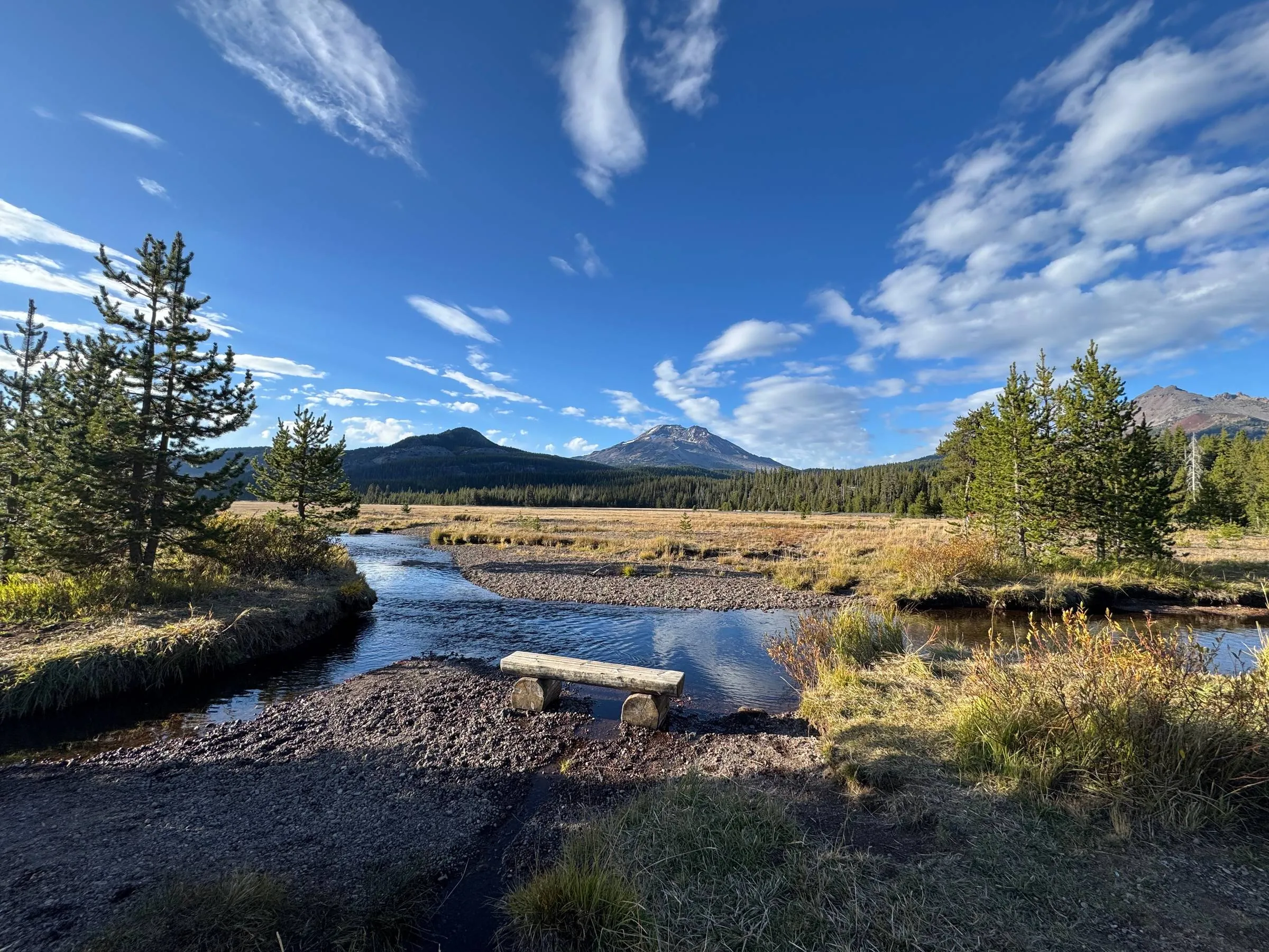 A creek winding through a golden autumn meadow with a log bench in the foreground, pine trees on both banks, and mountains under a blue sky with wispy clouds