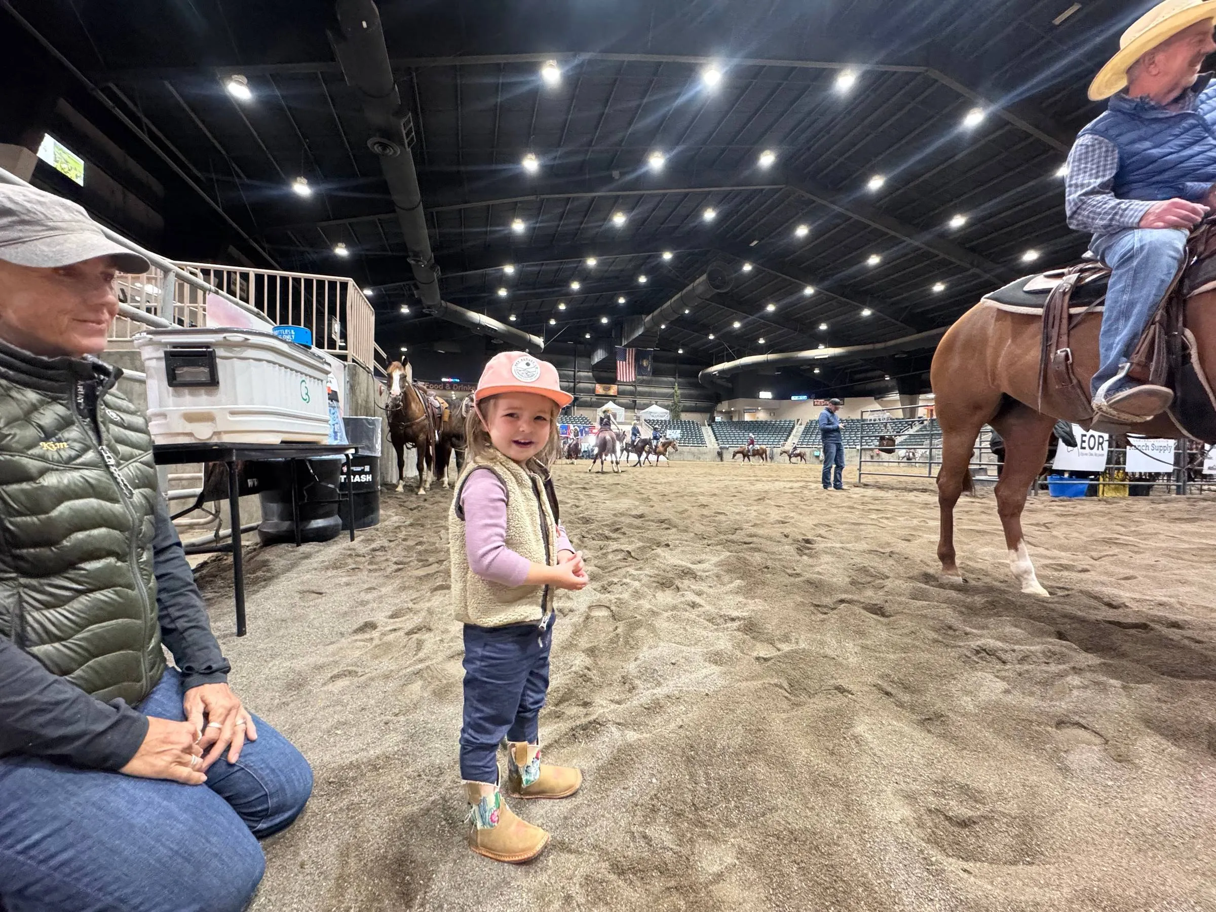 Ben’s daughter Parker in a pink ball cap and cowboy boots grinning inside an indoor arena while a rider on a chestnut horse waits nearby
