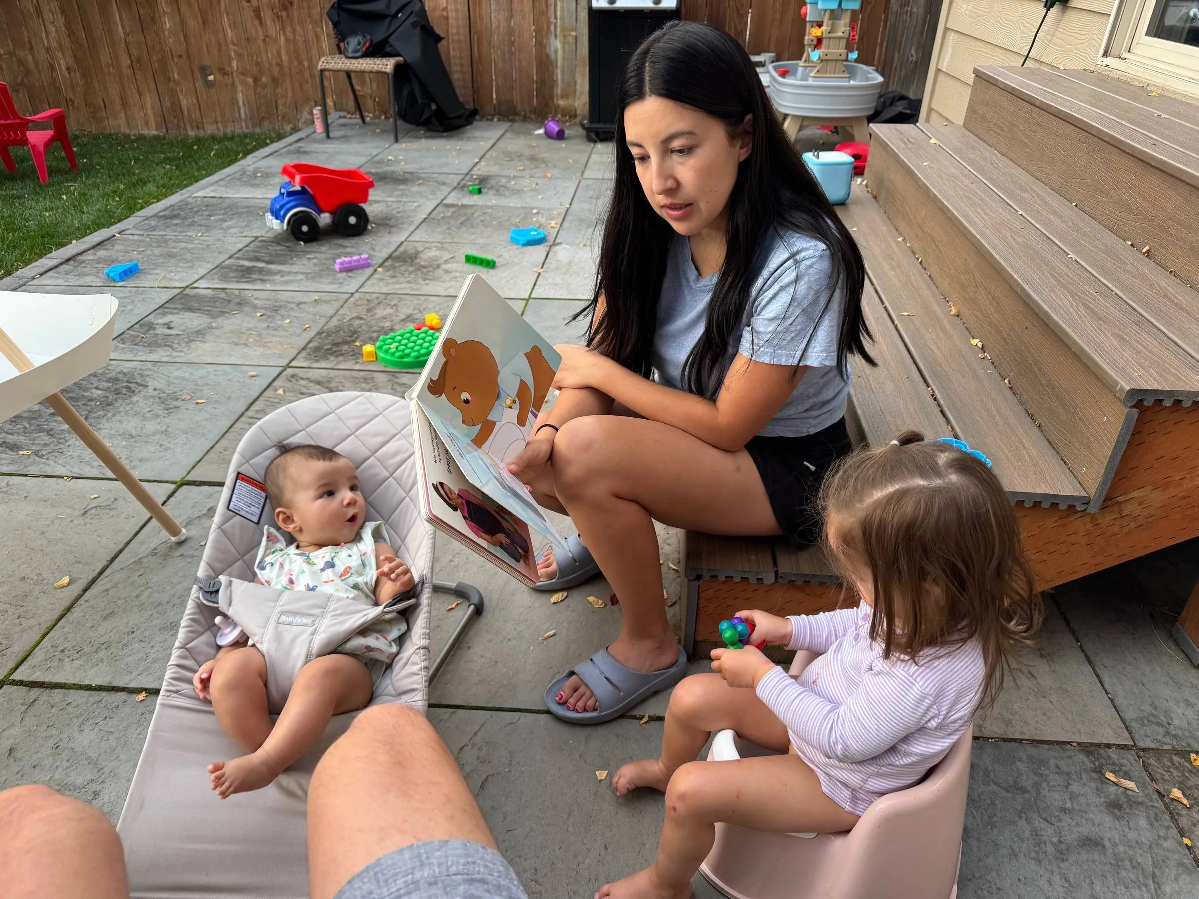 Ben’s wife sitting on the back deck steps reading a picture book to their baby in a bouncer and their toddler on a potty seat, with toddler toys scattered across the patio
