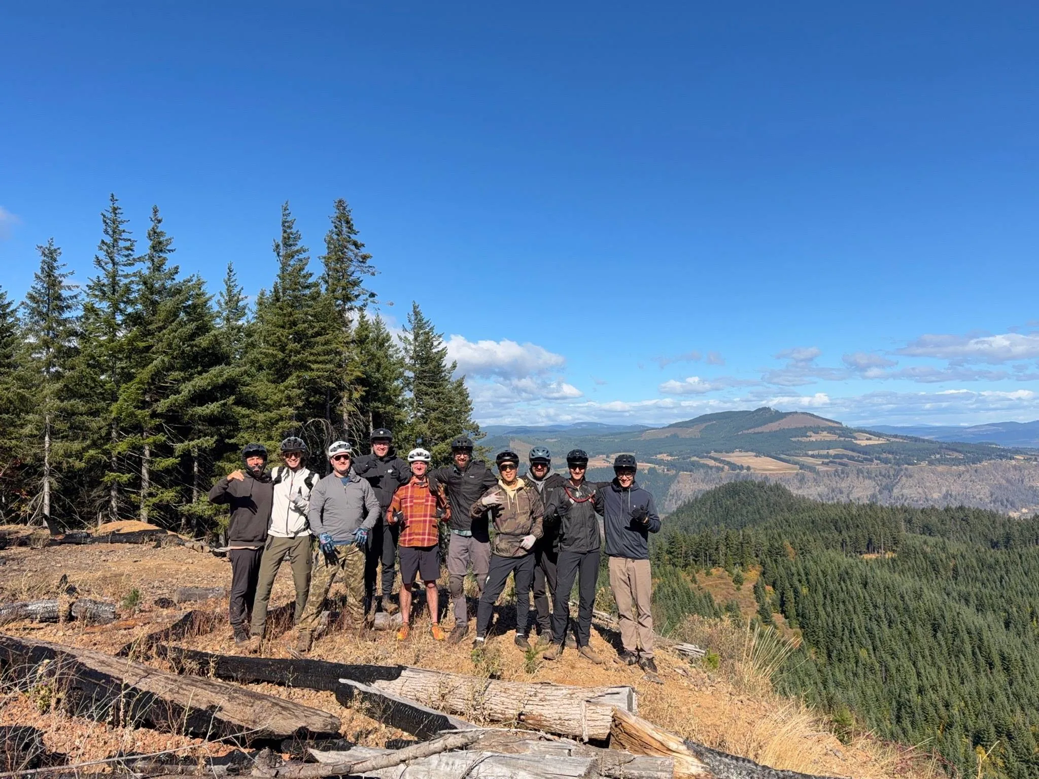 Ten mountain bikers in helmets posing on a ridge above Hood River, with evergreen trees and rolling hills behind them