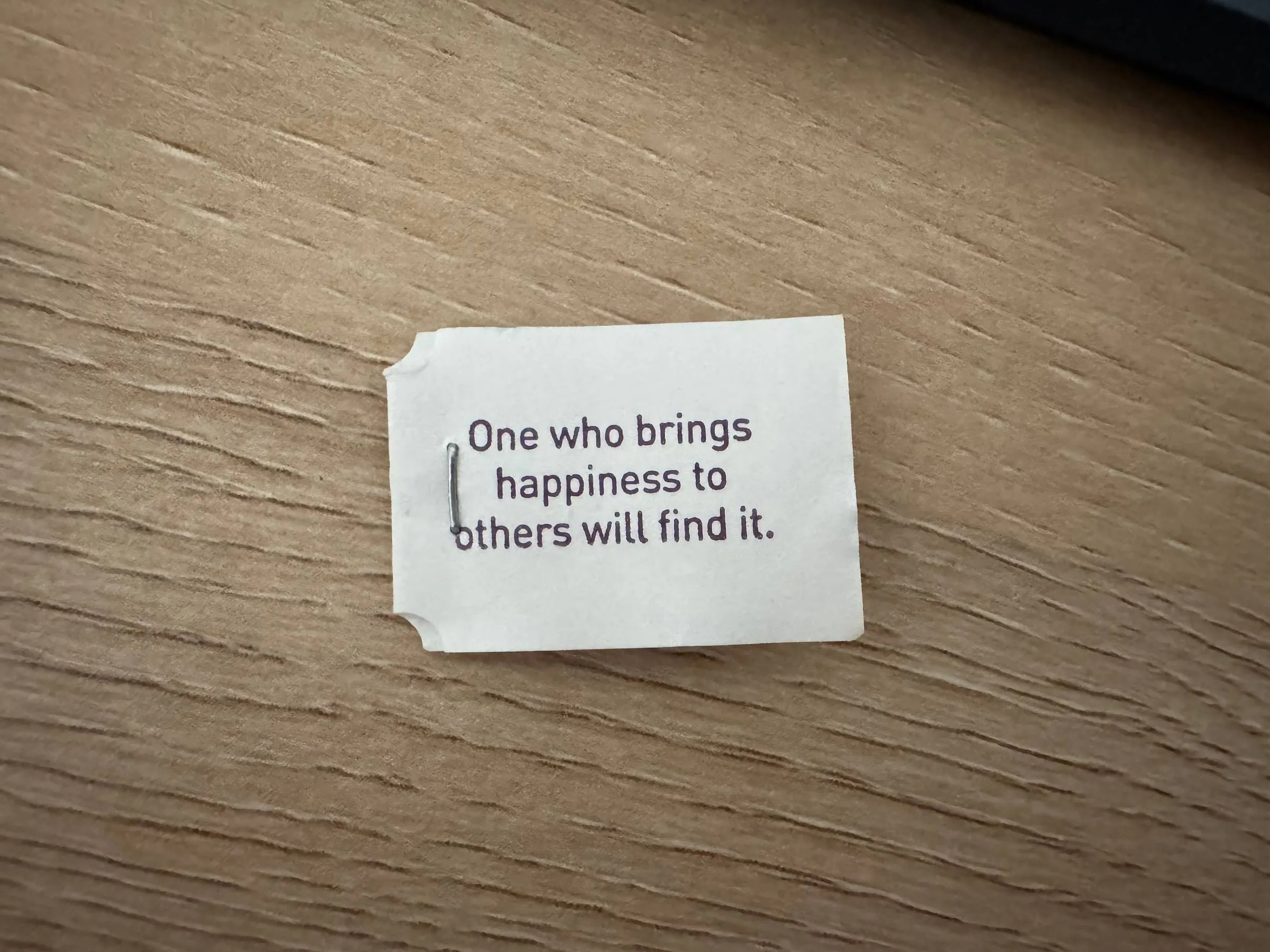 A small paper tea tag on a wooden table reading “One who brings happiness to others will find it.”