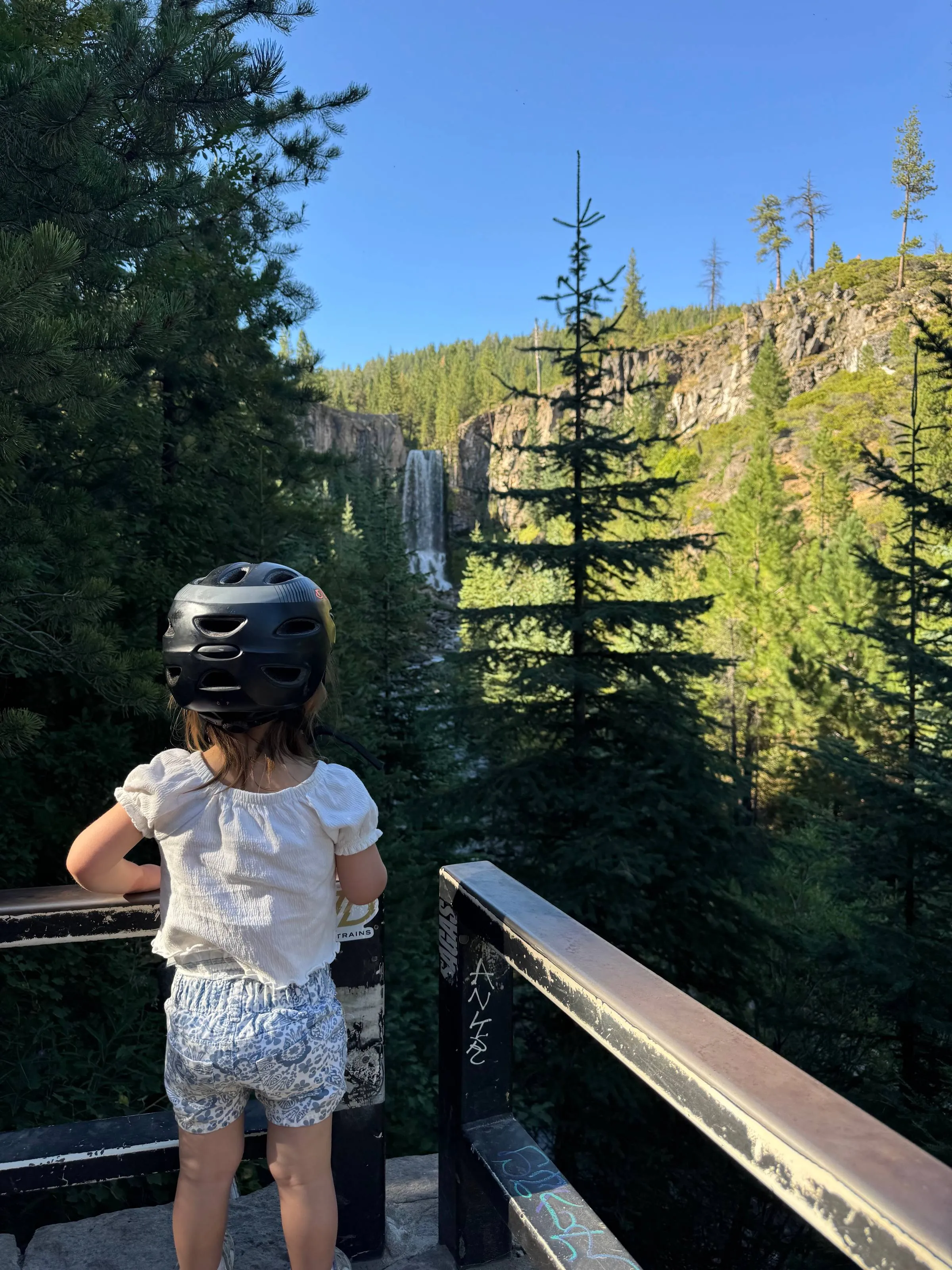 Ben’s daughter in a bike helmet leaning on a railing, looking out at Tumalo Falls and the pine-covered canyon behind it