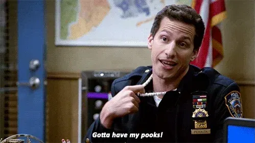 Brooklyn Nine-Nine GIF of Jake Peralta in a police uniform pointing at his puka shell necklace with the caption “Gotta have my pooks!”