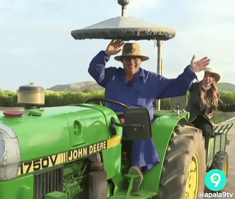 A man in a wide-brimmed hat waving with both arms while driving a green John Deere tractor down a country road